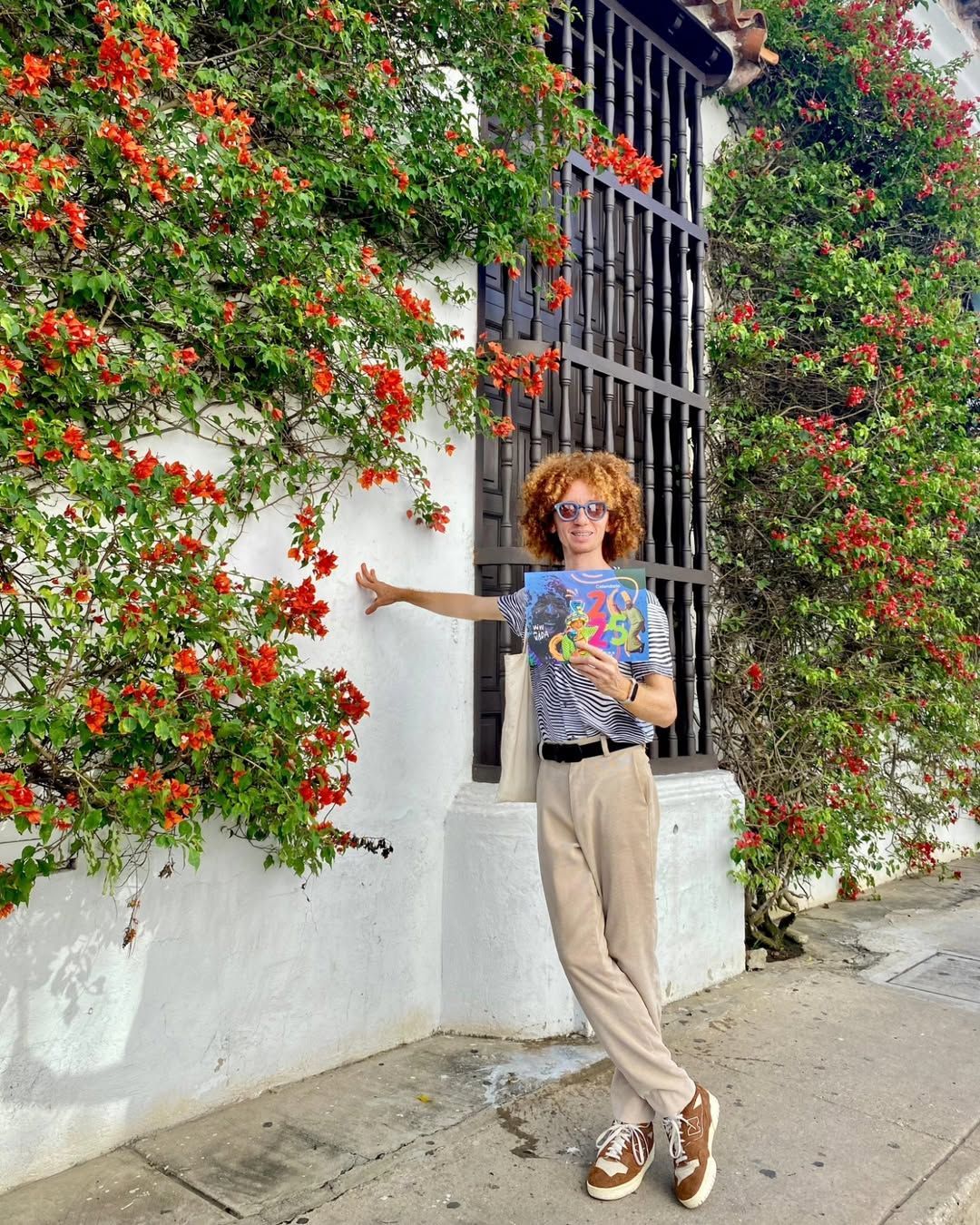 Persona posando junto a una pared blanca cubierta de enredaderas de flores rojas, sosteniendo una colorida lámina artística.