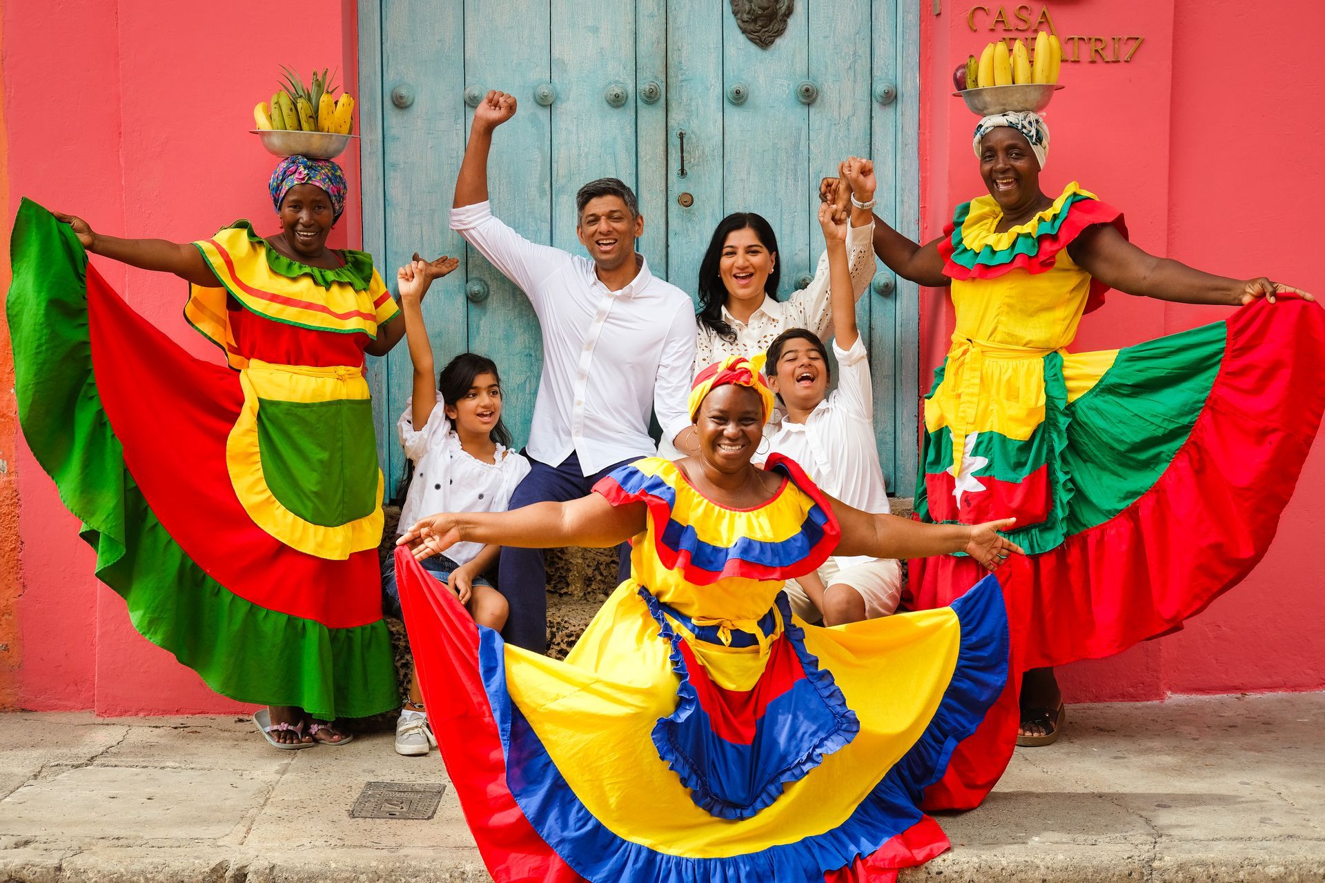 Bailarines con coloridos trajes tradicionales posan frente a una pared rosa, con los brazos extendidos.