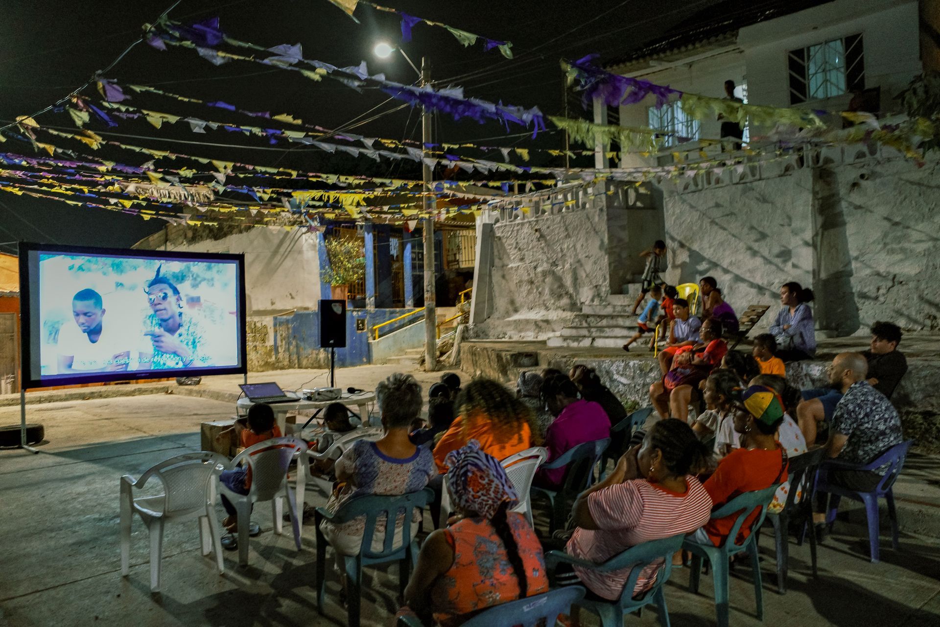 Proyección nocturna de cine al aire libre con gente sentada en sillas bajo guirnaldas de luces.