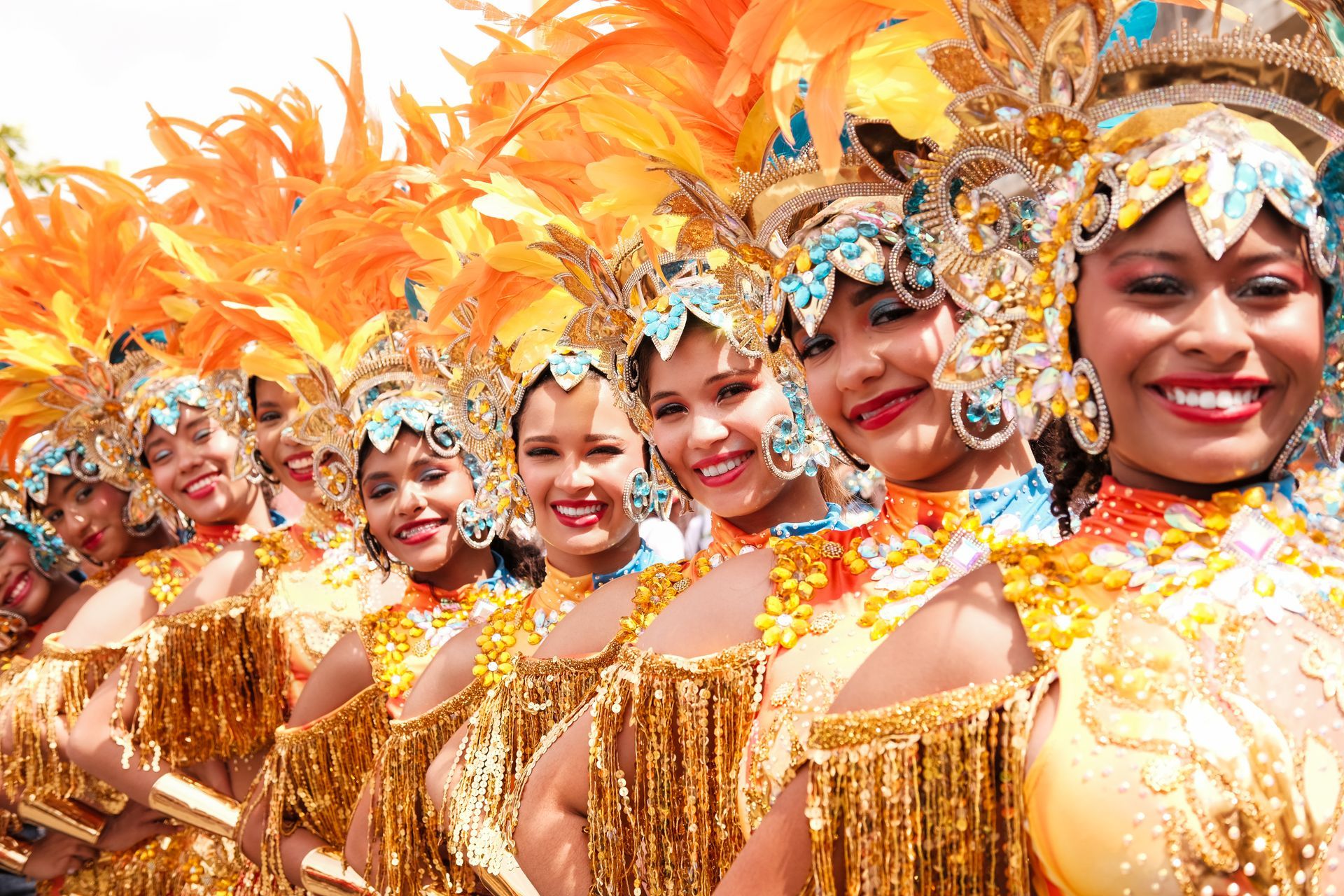 Fila de artistas con trajes dorados y tocados de plumas sonriendo durante un vibrante desfile.