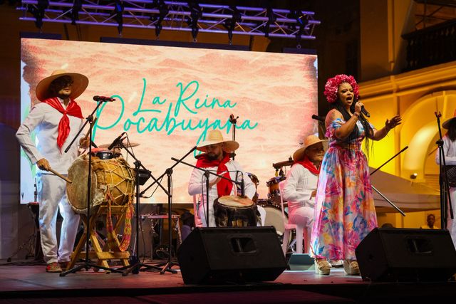 Banda en vivo actuando en el escenario con batería, guitarra y cantante con coloridos atuendos bajo luces cálidas.