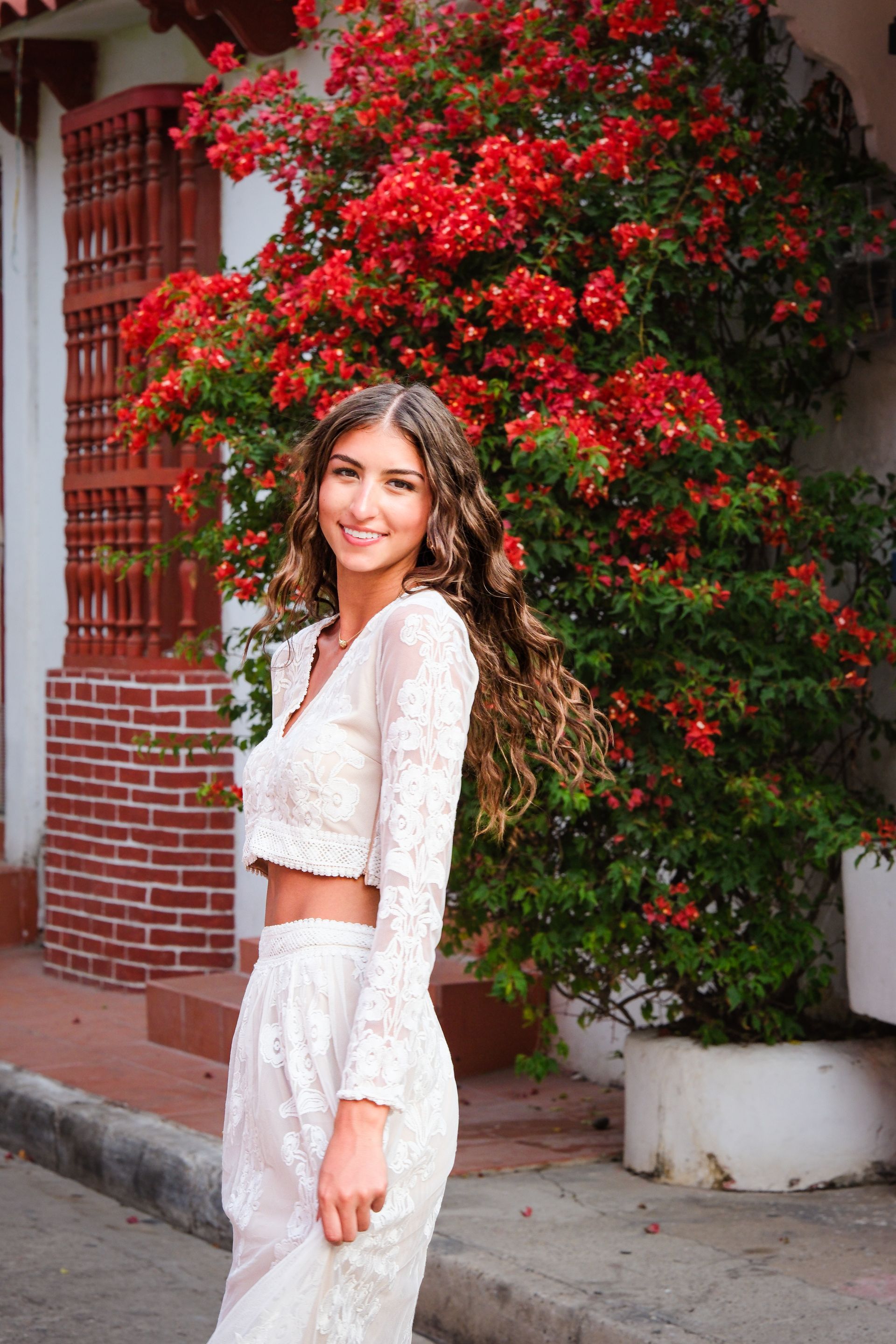 Mujer vestida de blanco de pie junto a un muro de ladrillos cubierto de flores de color rojo brillante.