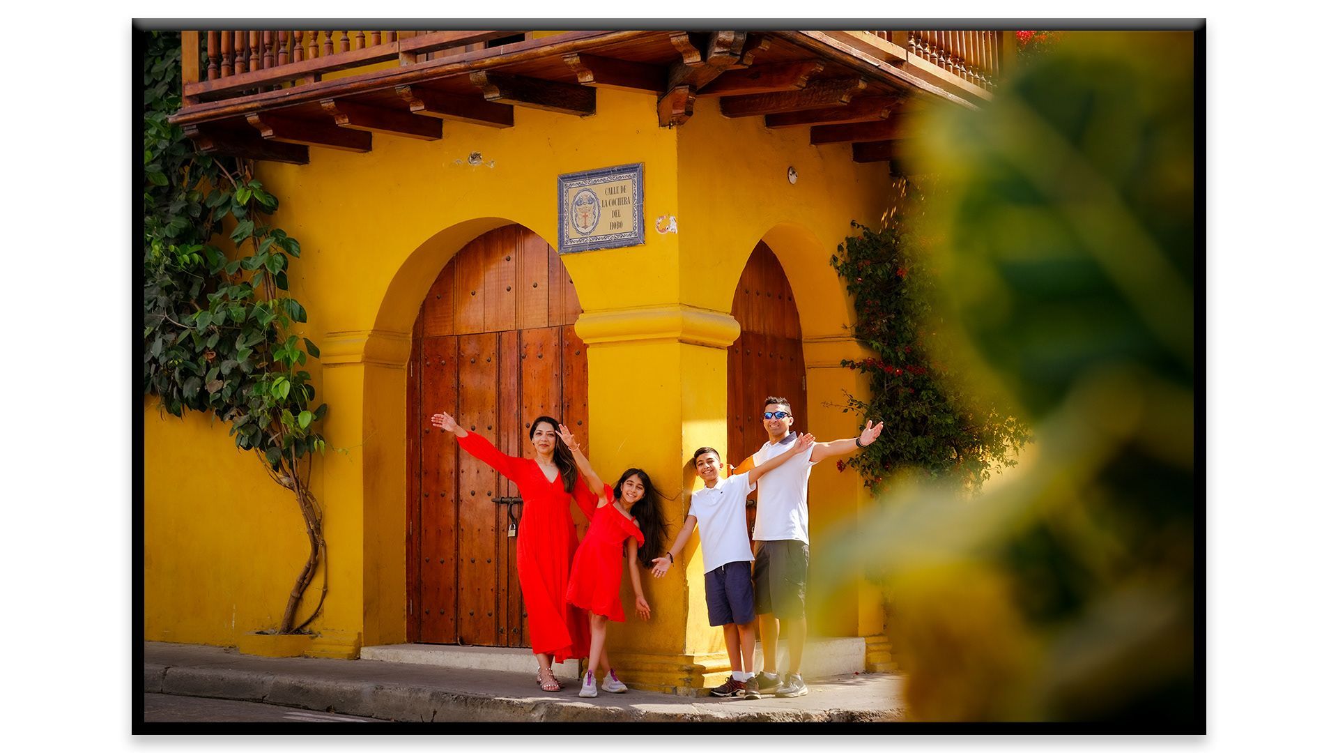 Tres personas posan frente a una puerta arqueada amarilla, una de ellas con un vestido rojo, en un patio soleado.