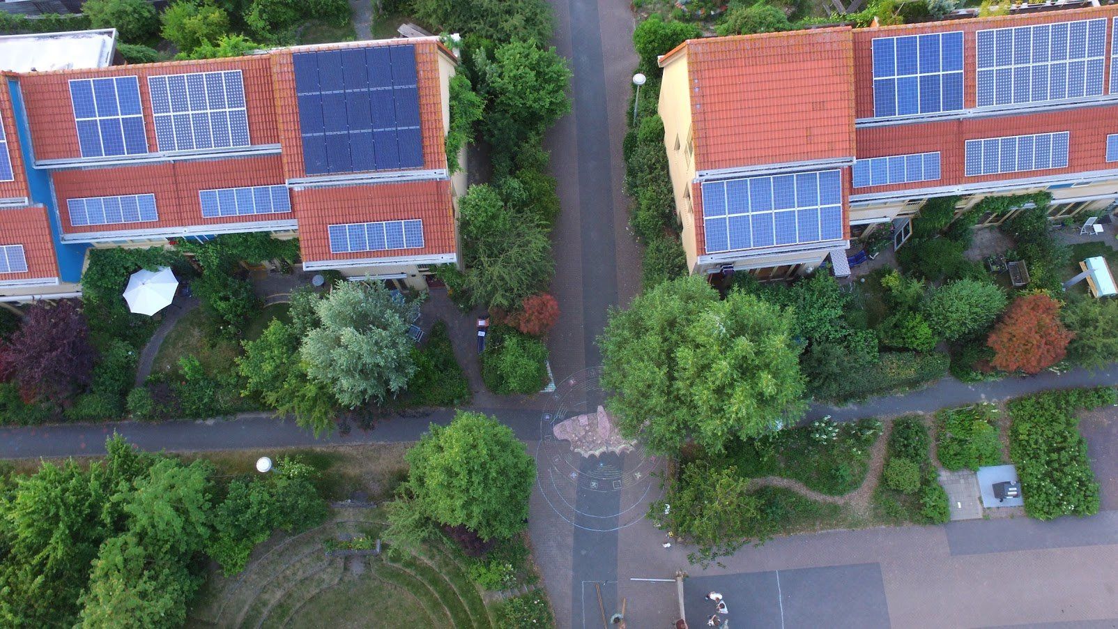 Daken van woningen met zonnepanelen, rode dakpannen, met uitzicht op een straat met bomen, groen en een tuin.