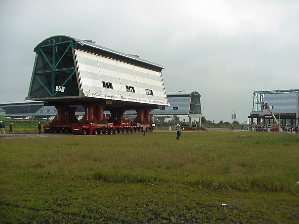 Drie grote, zilverkleurige en groene industriële constructies op rode trailers, op een grasveld; mensen in de buurt.