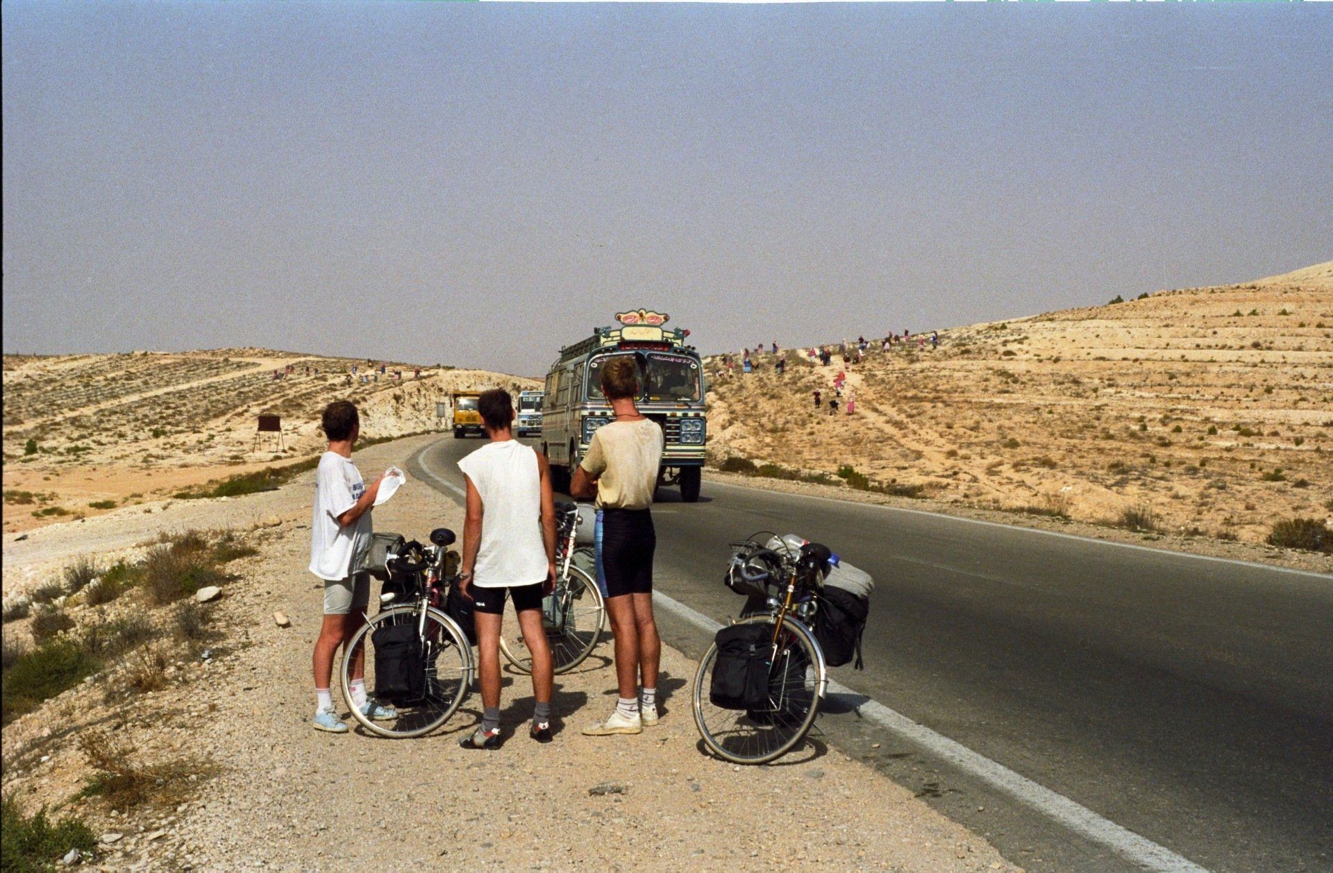 Drie fietsers staan ​​langs de weg in de woestijn en kijken naar een bus. De fietsen hebben fietstassen.