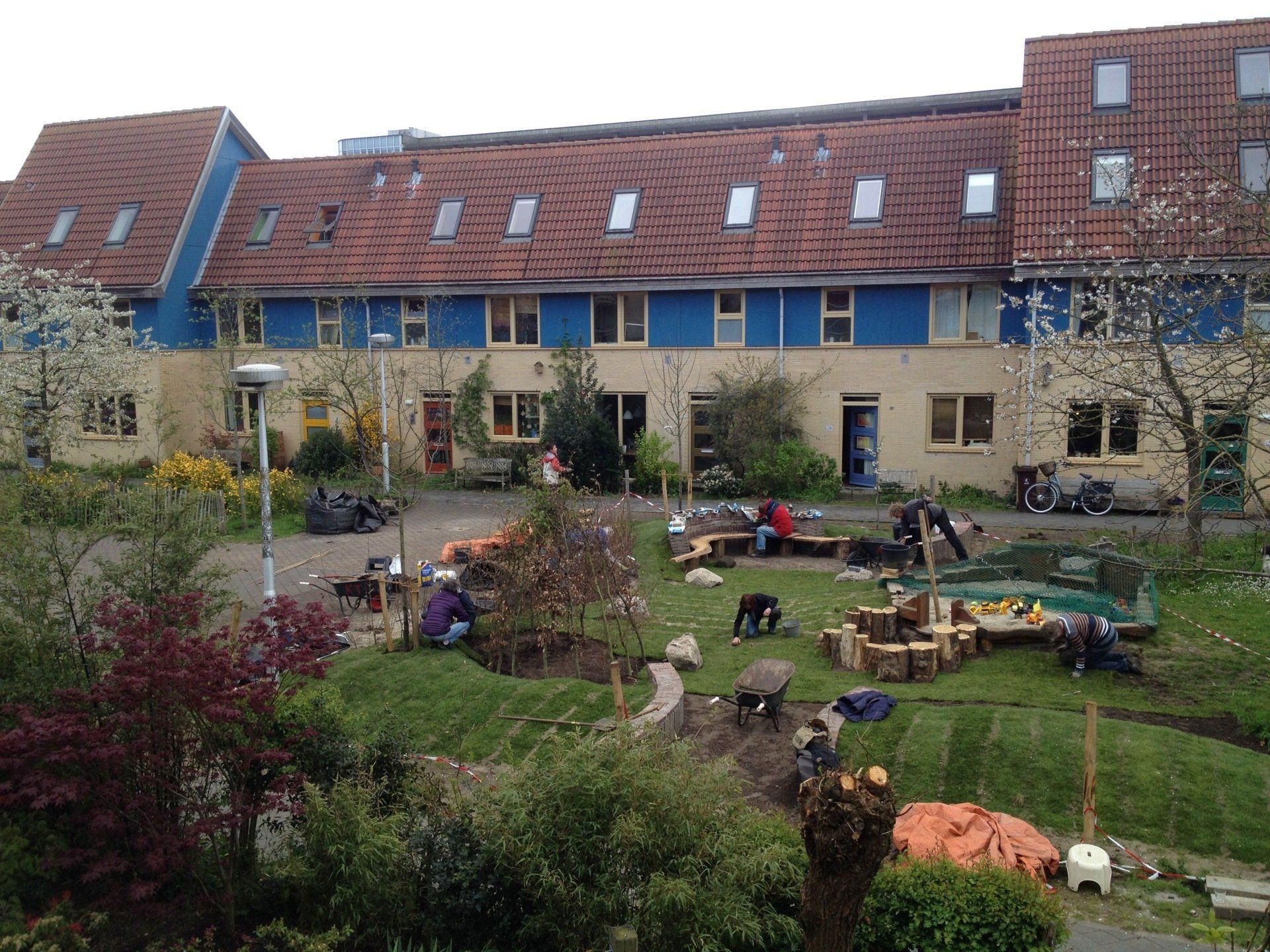 Rijtjeshuizen met blauwe kozijnen en rode pannendaken; mensen die in de voortuin tuinieren.