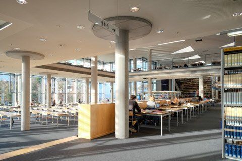 Licht en modern bibliotheekinterieur met zuilen, tafels, stoelen en boekenkasten. Zonlicht komt door grote ramen naar binnen.