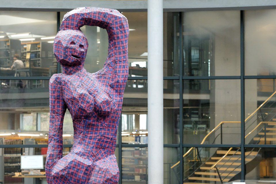 Kunstculptuur van een vrouw met opgeheven arm, staande in een entreehal van een bibliotheek.