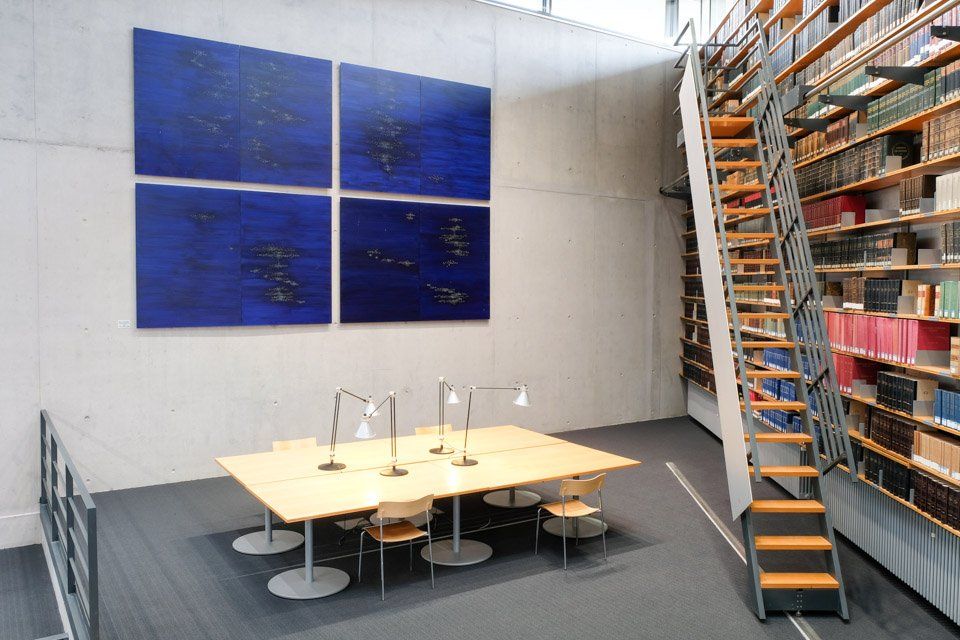 Modern bibliotheekinterieur met leestafel, boekenkasten en een blauw kunstwerk aan de muur.