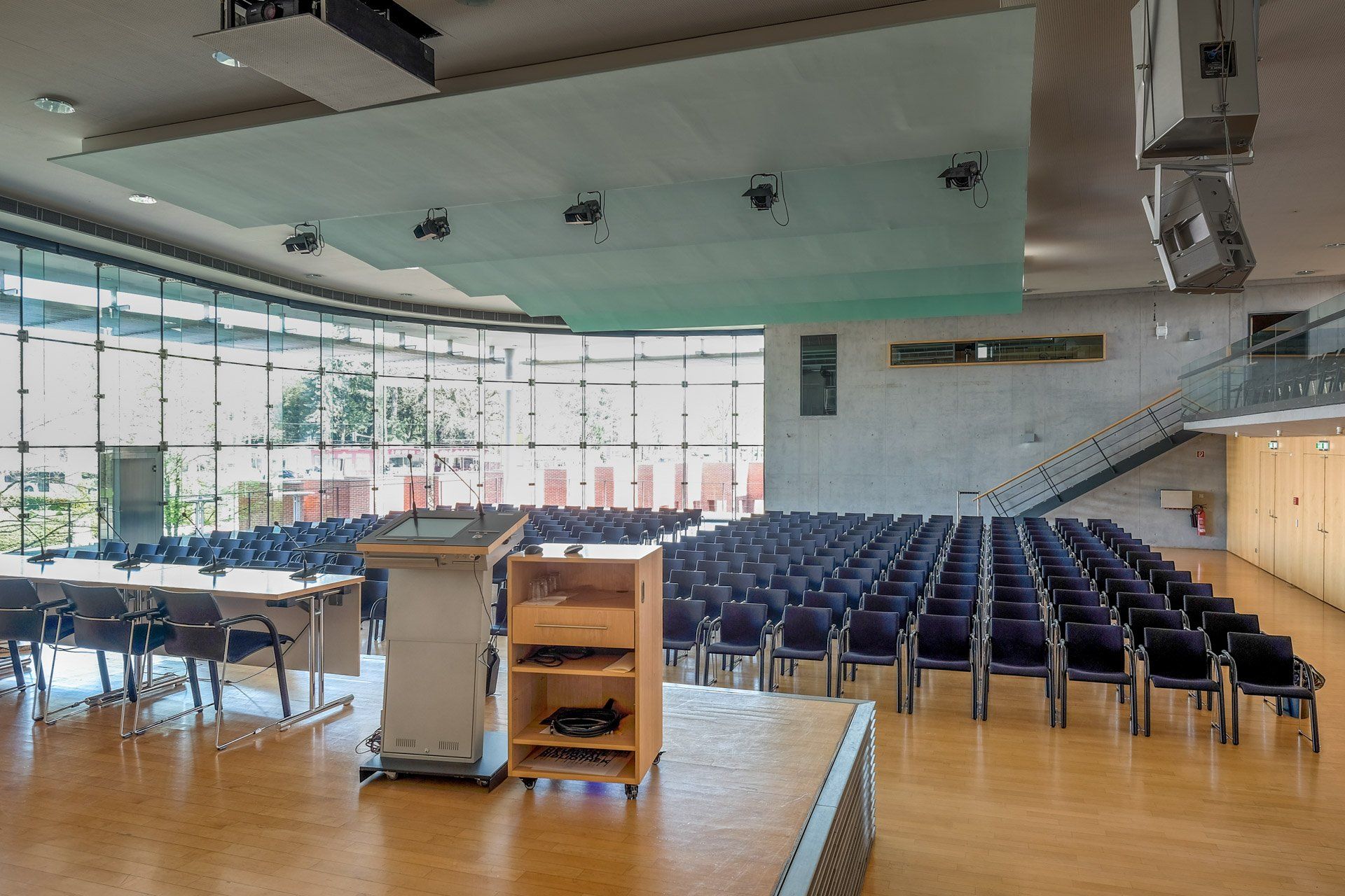 Een lege zaal met rijen blauwe stoelen tegenover een podium. Grote ramen aan de linkerkant en een trap aan de rechterkant.
