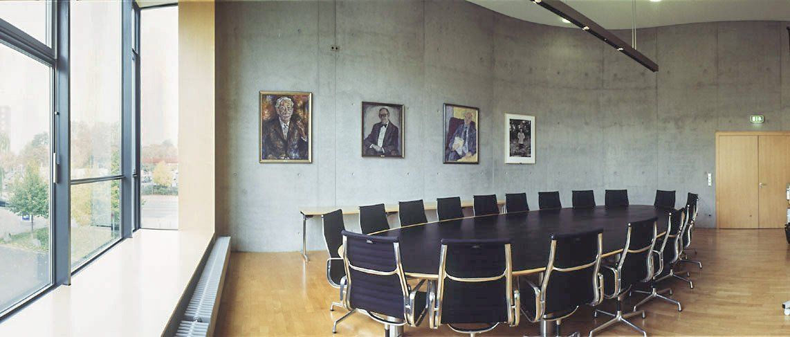 Een moderne vergaderruimte met een grote ovale tafel en stoelen, en aan de muur hangen portretten.