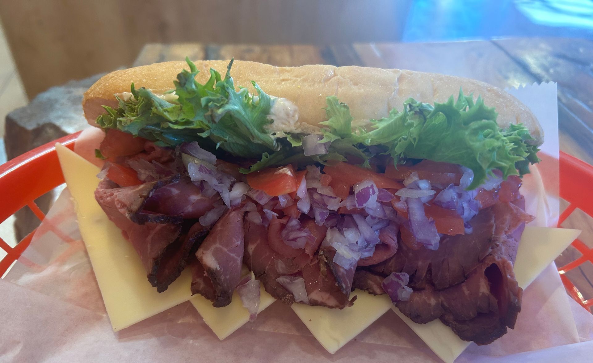 A sub sandwich with roast beef , lettuce , tomatoes and cheese in a red basket.