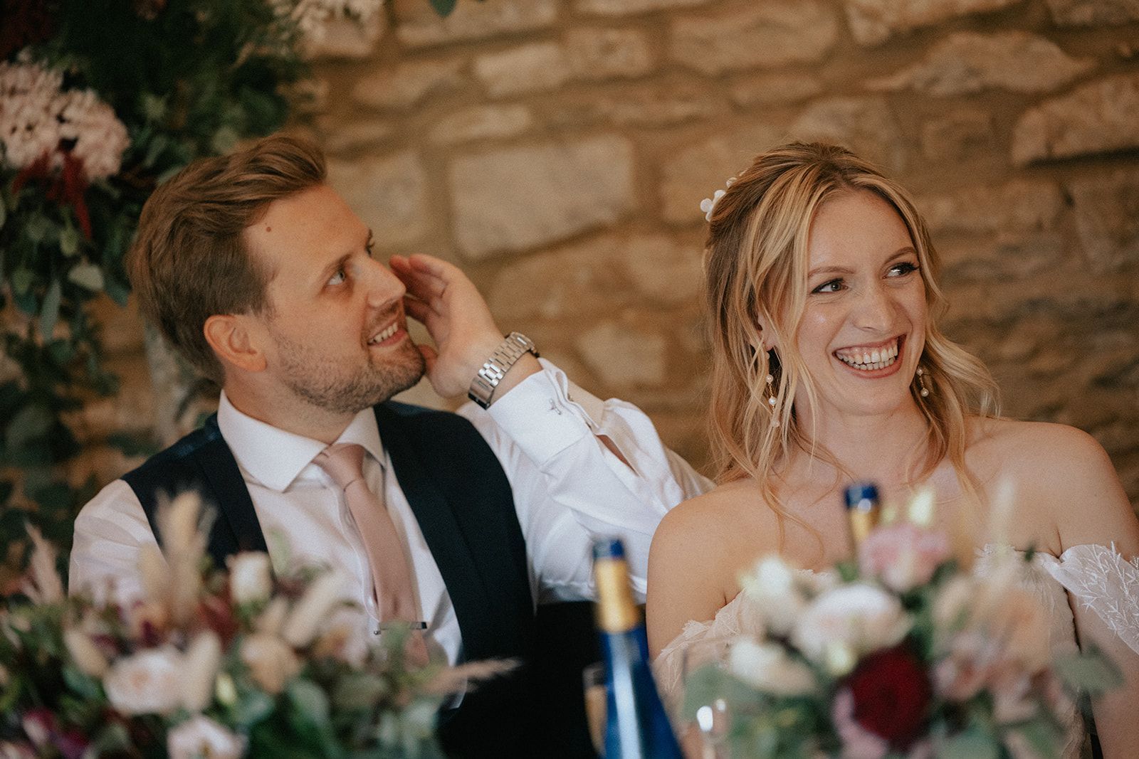 A bride and groom are sitting at a table with a bottle of wine.