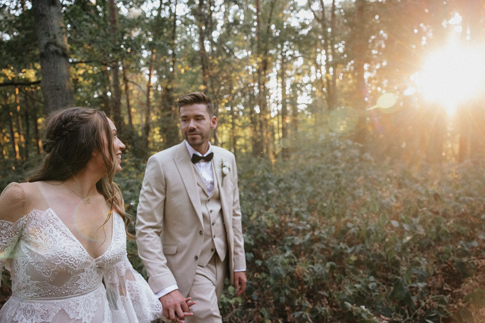 A bride and groom are walking through the woods holding hands.