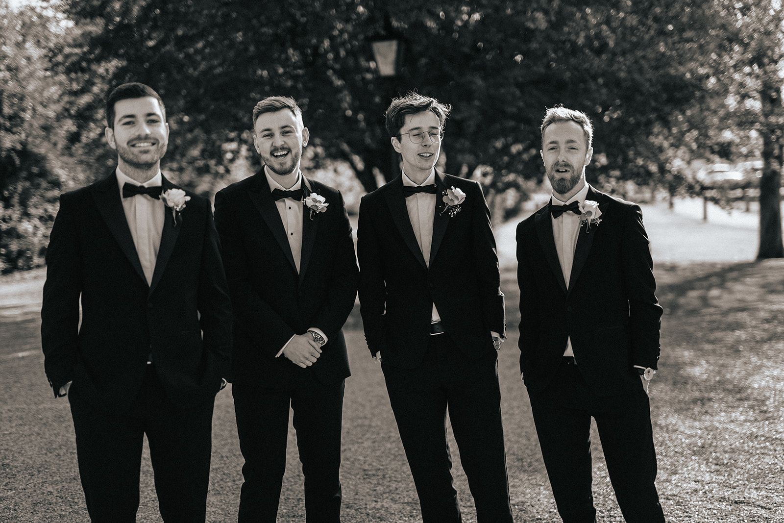 A group of men in tuxedos and bow ties are standing next to each other in a park.