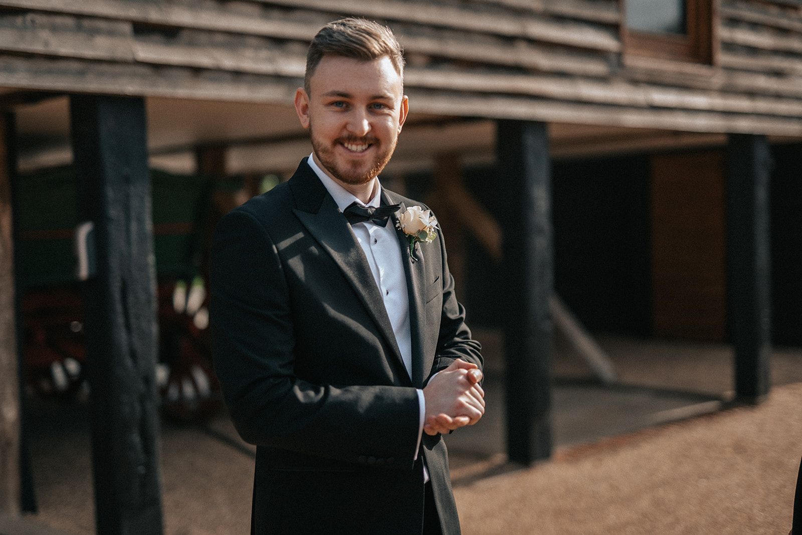 A man in a tuxedo and bow tie is standing in front of a wooden building.