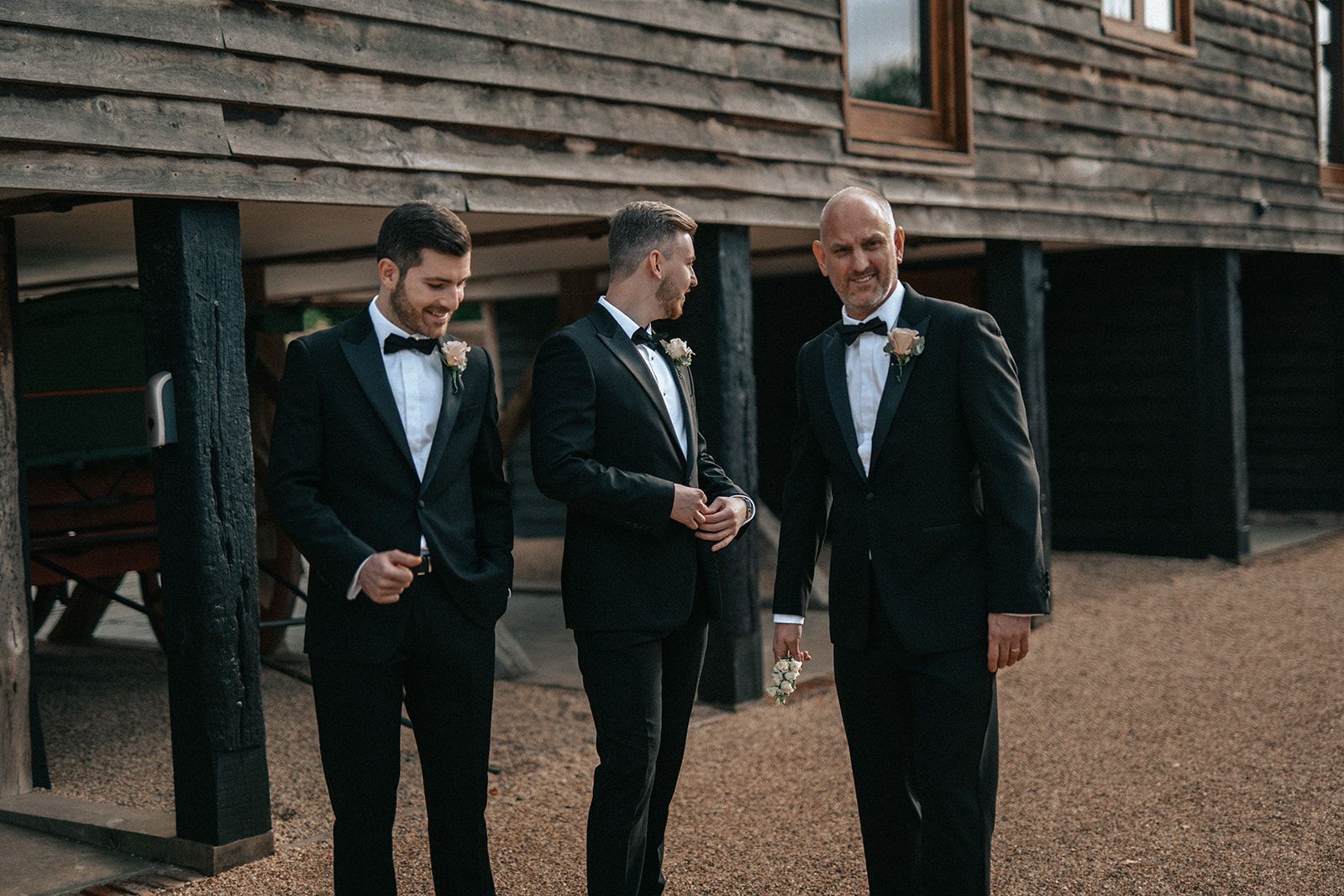 Three men in tuxedos are standing in front of a wooden building.