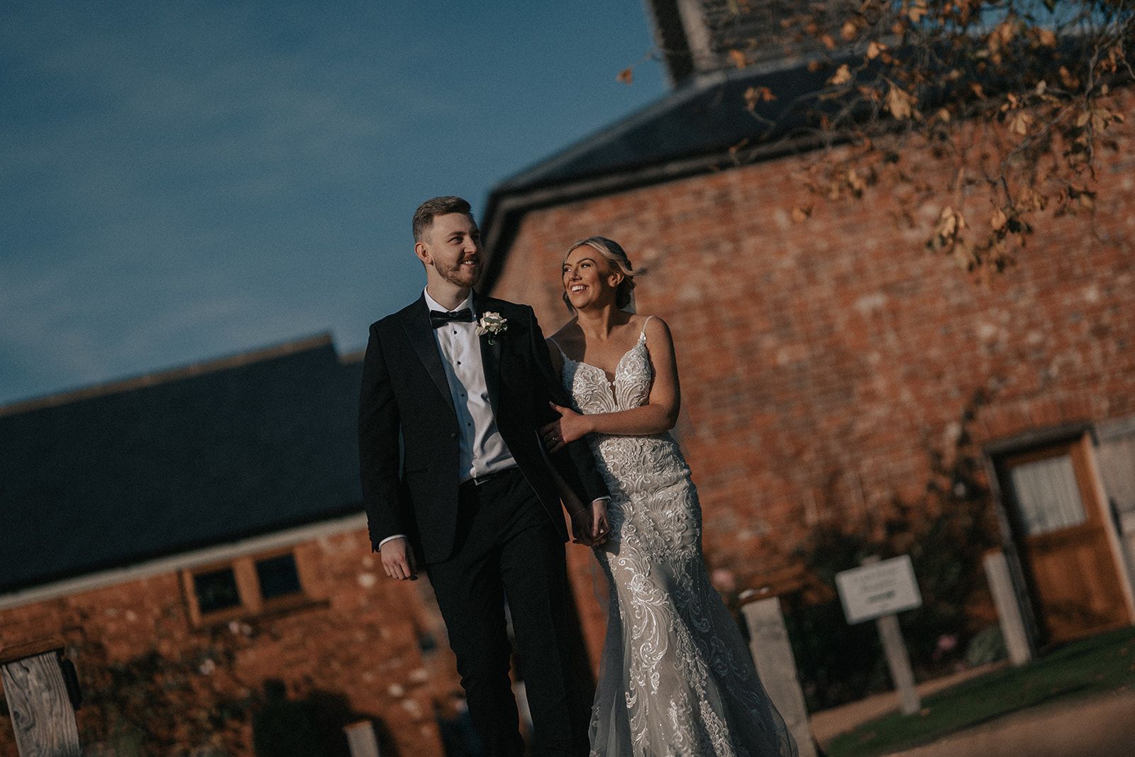 A bride and groom are walking in front of a brick building holding hands.
