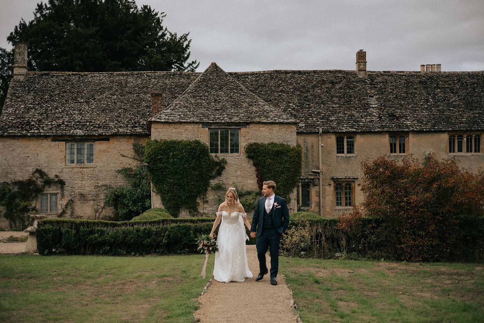A bride and groom are walking down a path in front of a large stone building.