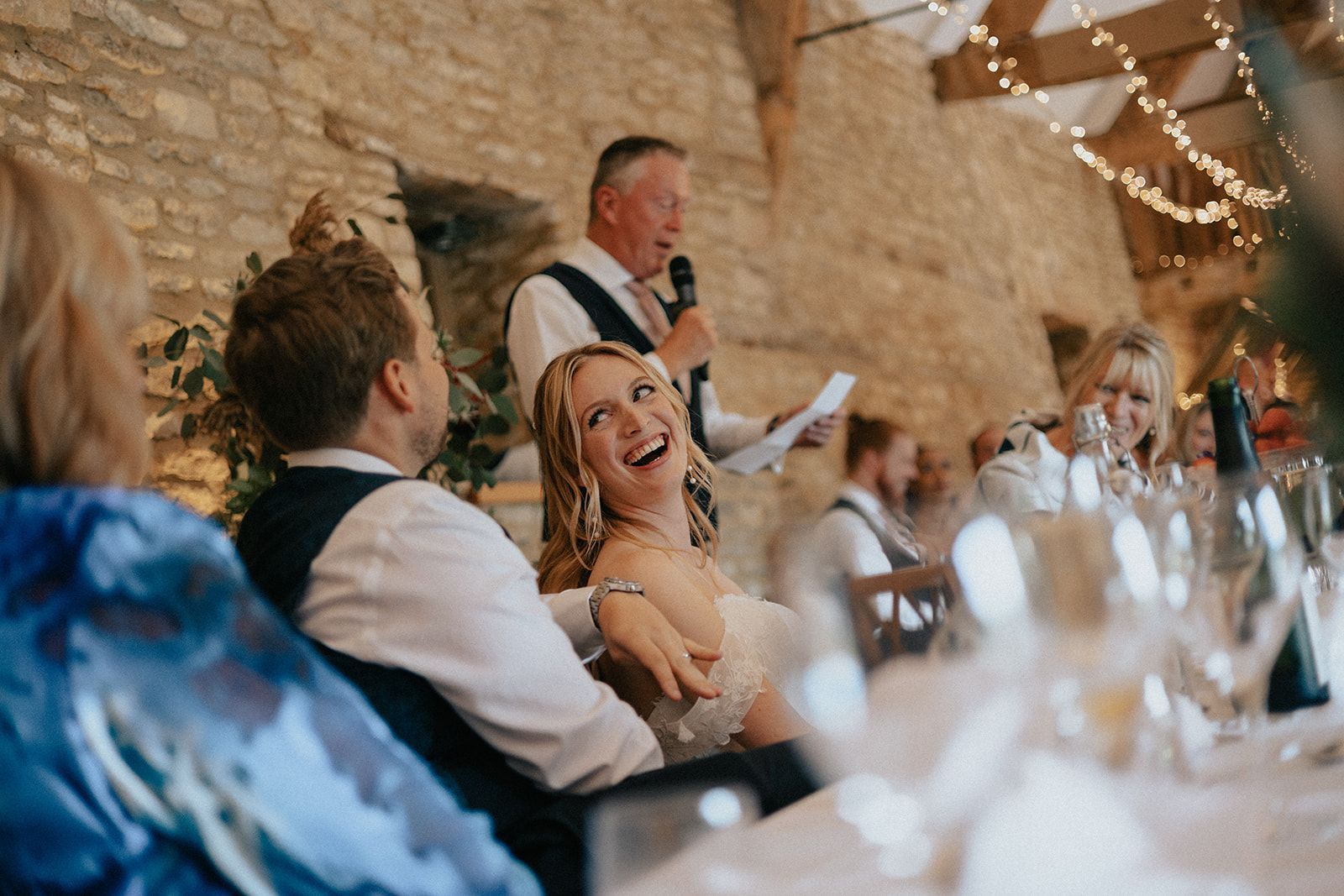 A bride and groom are laughing while a man holds a microphone at a wedding reception.