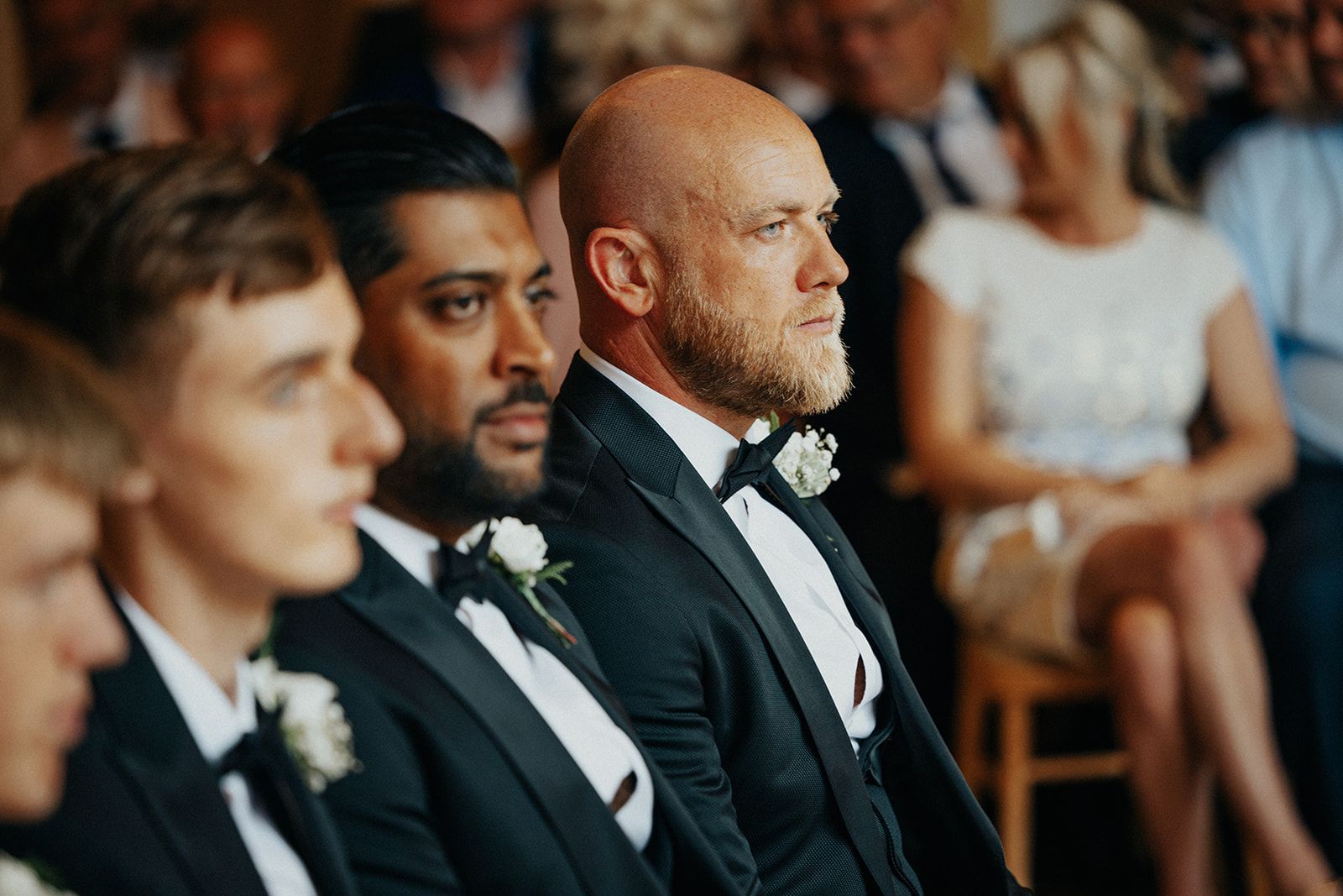 A group of men in tuxedos are sitting in a row at a wedding ceremony.