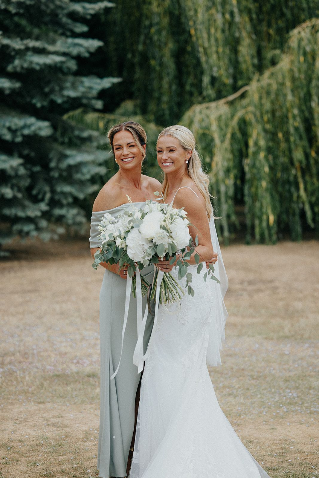 A bride and her bridesmaid are standing next to each other in a field holding flowers.