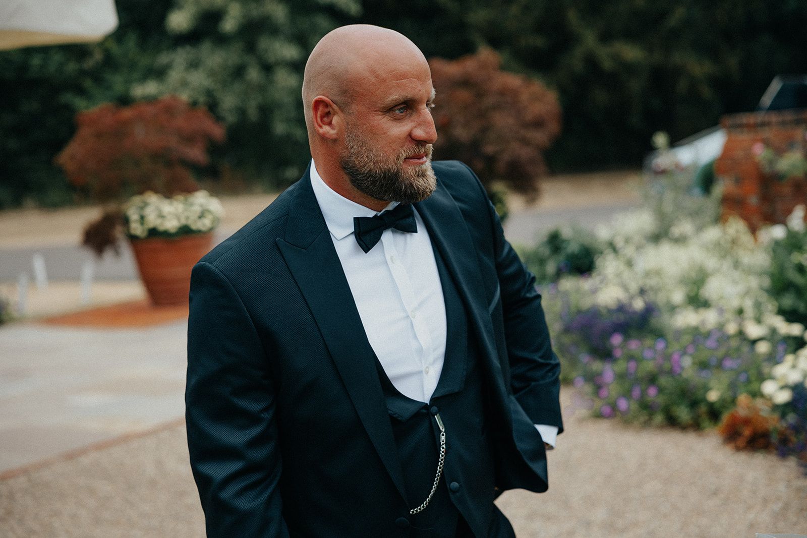 A man in a suit and bow tie is standing in front of a garden.
