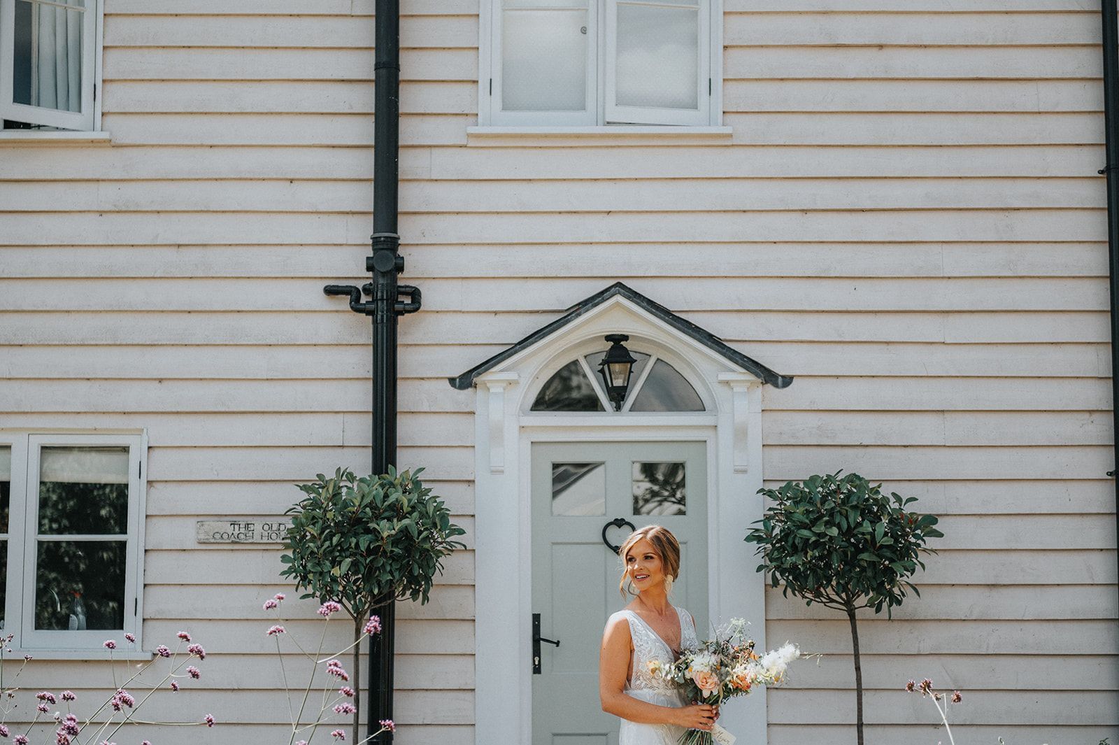 A bride is standing in front of a white house holding a bouquet of flowers.