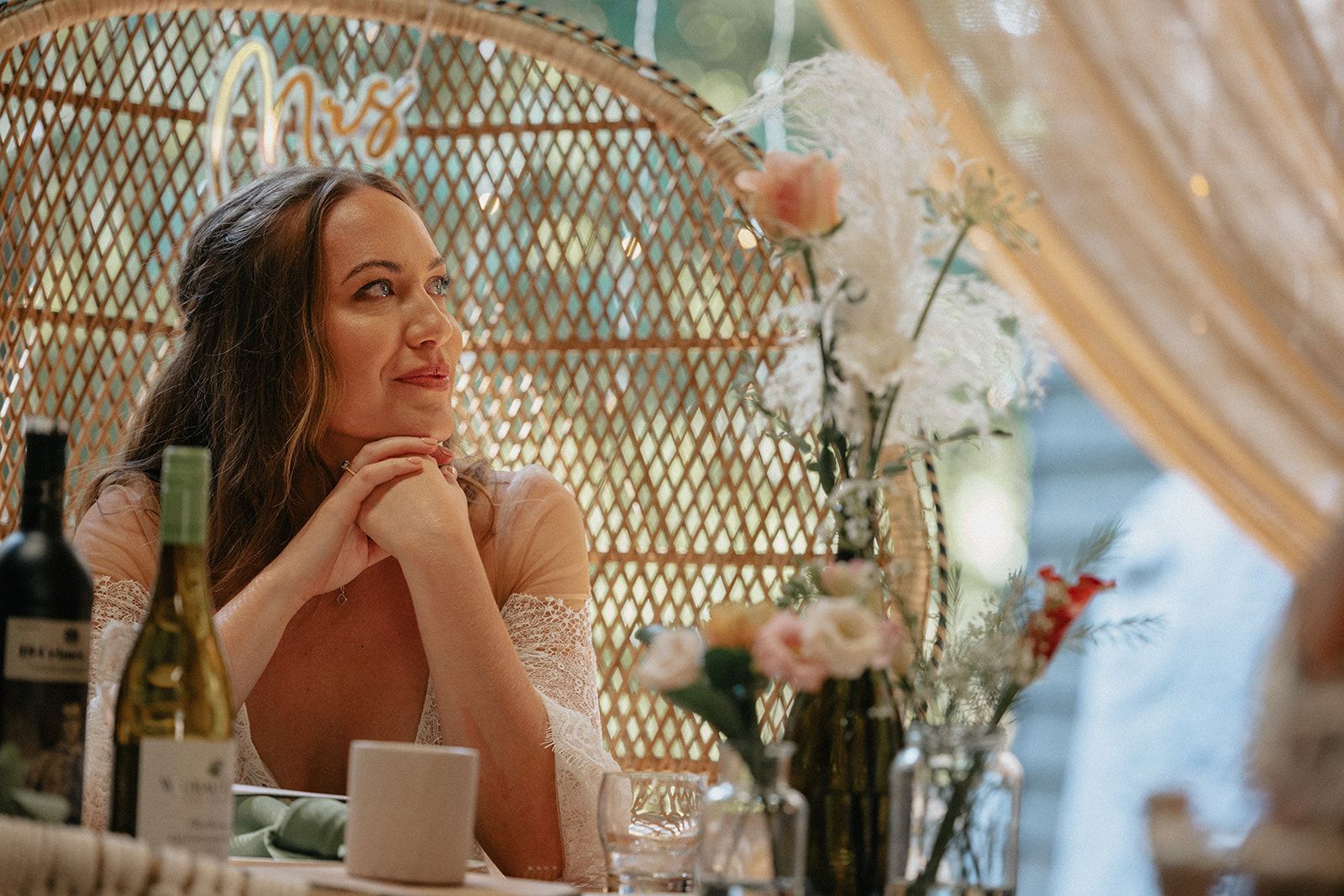 A woman is sitting in a peacock chair at a table with a bottle of wine.