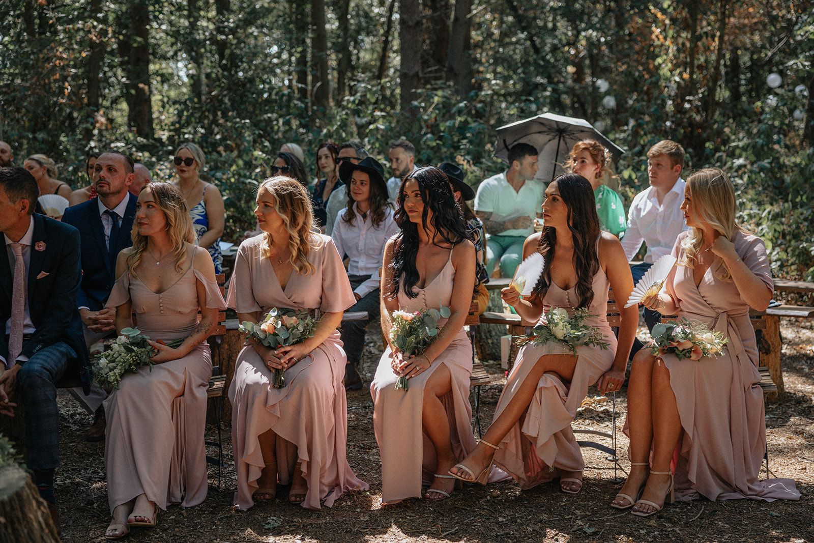 A group of bridesmaids are sitting in a row at a wedding ceremony in the woods.
