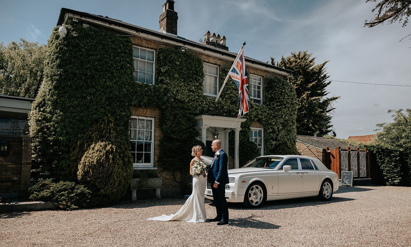 A bride and groom are standing in front of a white car in front of a house.