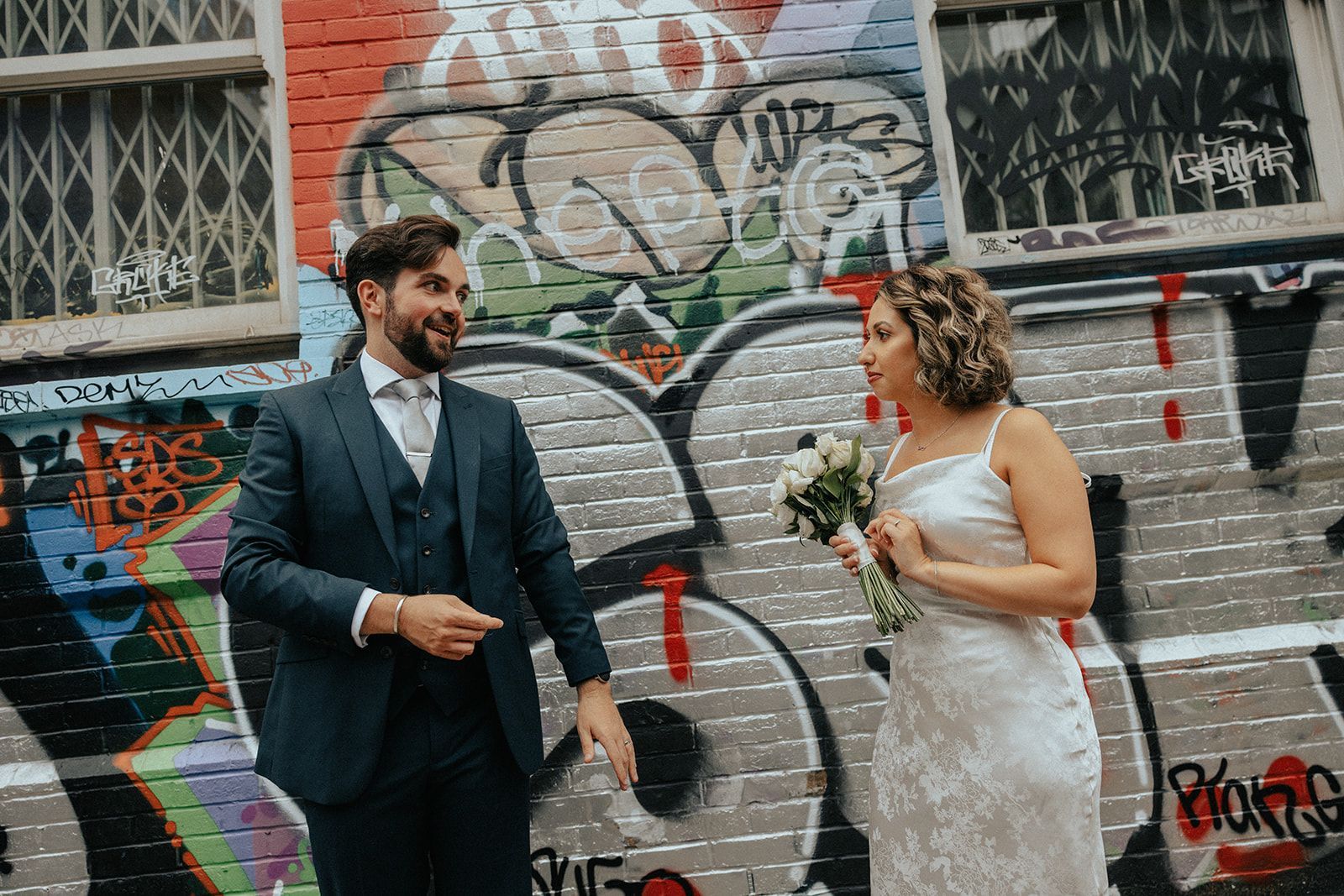 A bride and groom are standing in front of a graffiti covered brick wall.