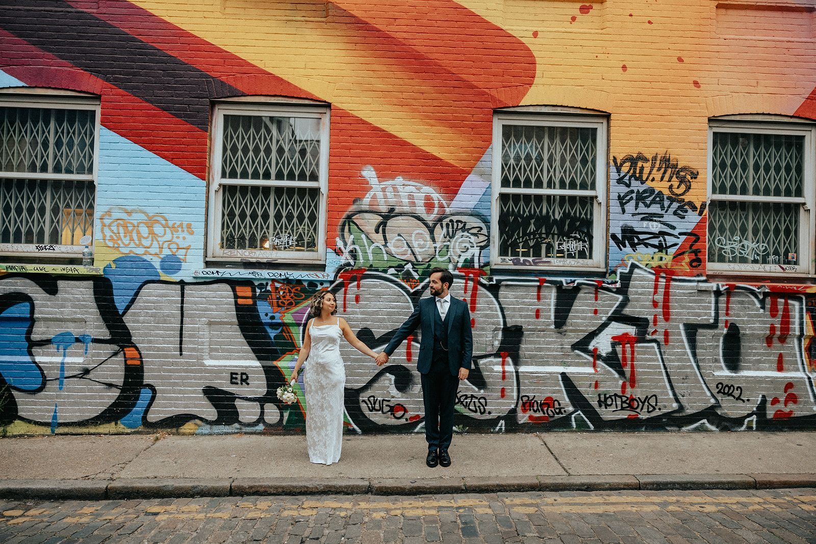 A bride and groom are holding hands in front of a graffiti covered building.