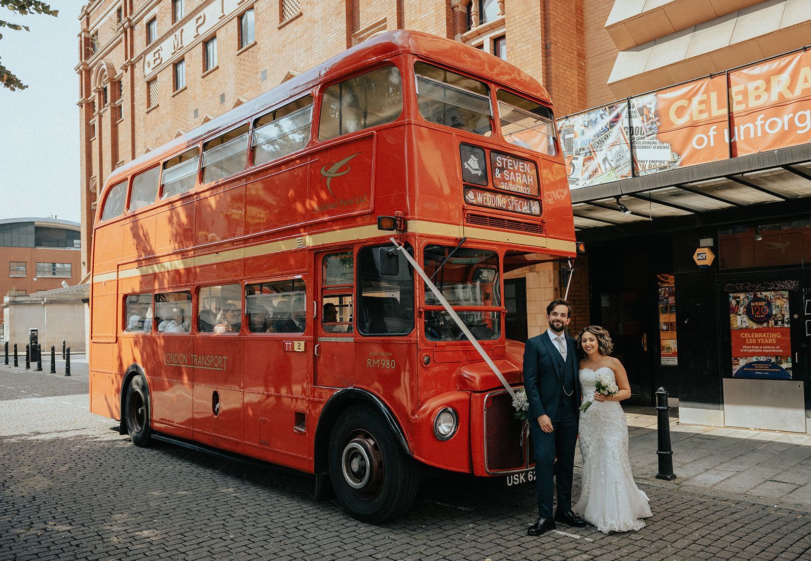 A bride and groom are standing in front of a red double decker bus.
