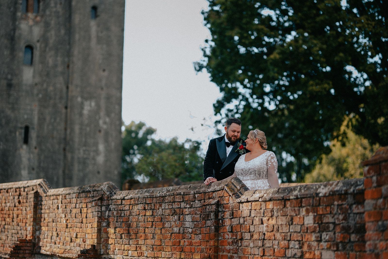 A bride and groom are sitting on a brick wall in front of a castle.