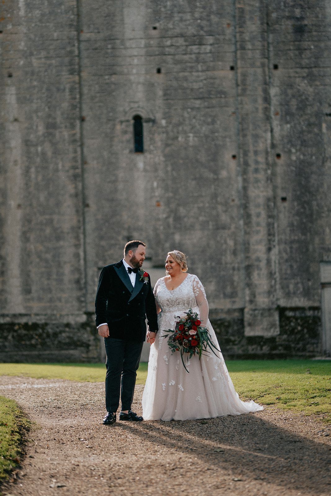 A bride and groom are walking down a dirt path holding hands in front of a building.