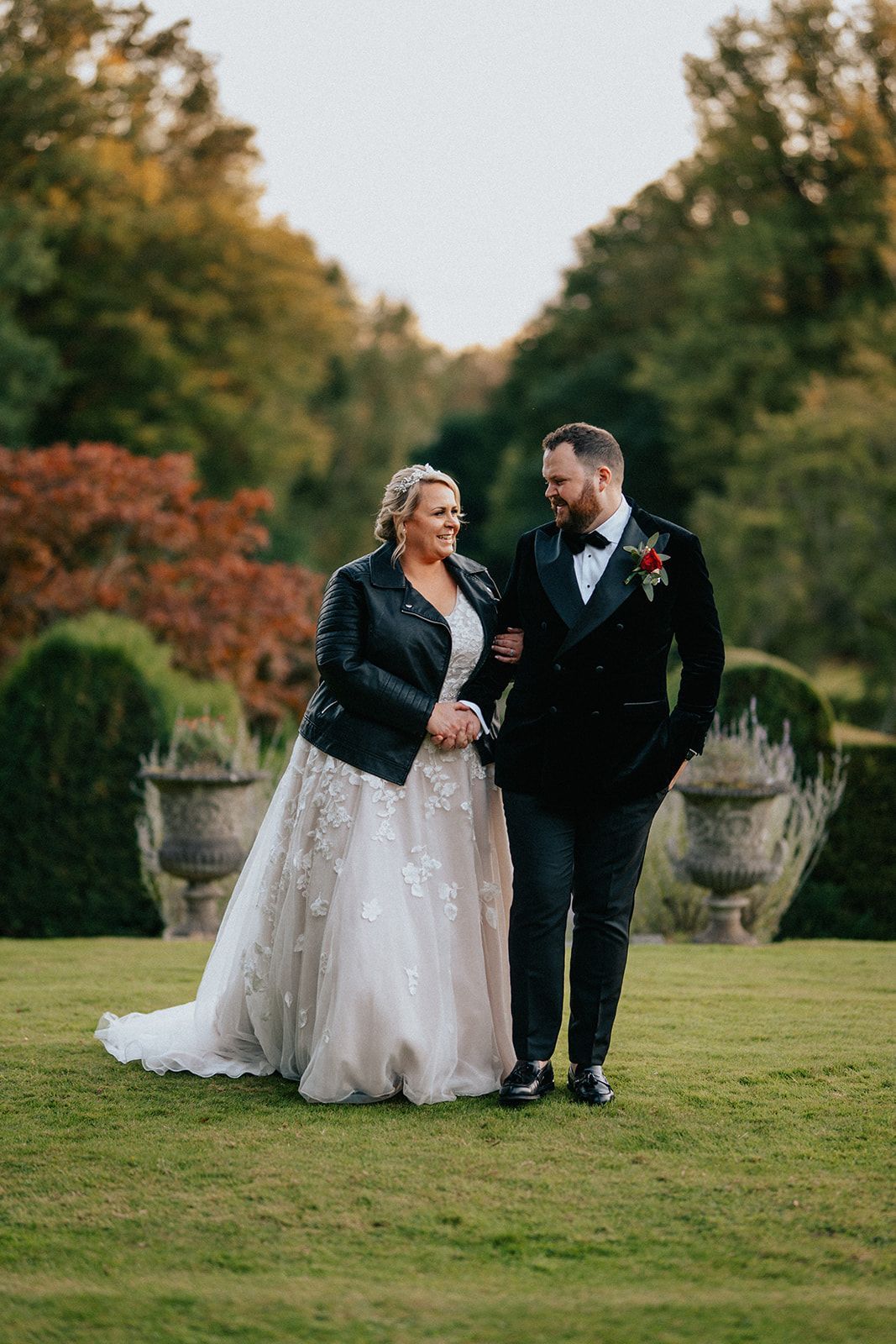 A bride and groom are walking on a lush green field holding hands.