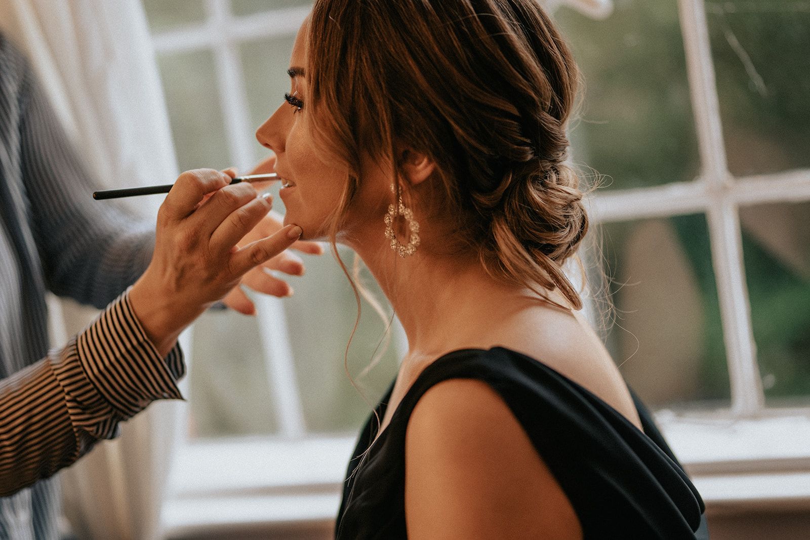 A woman is getting her makeup done by a makeup artist in front of a window.