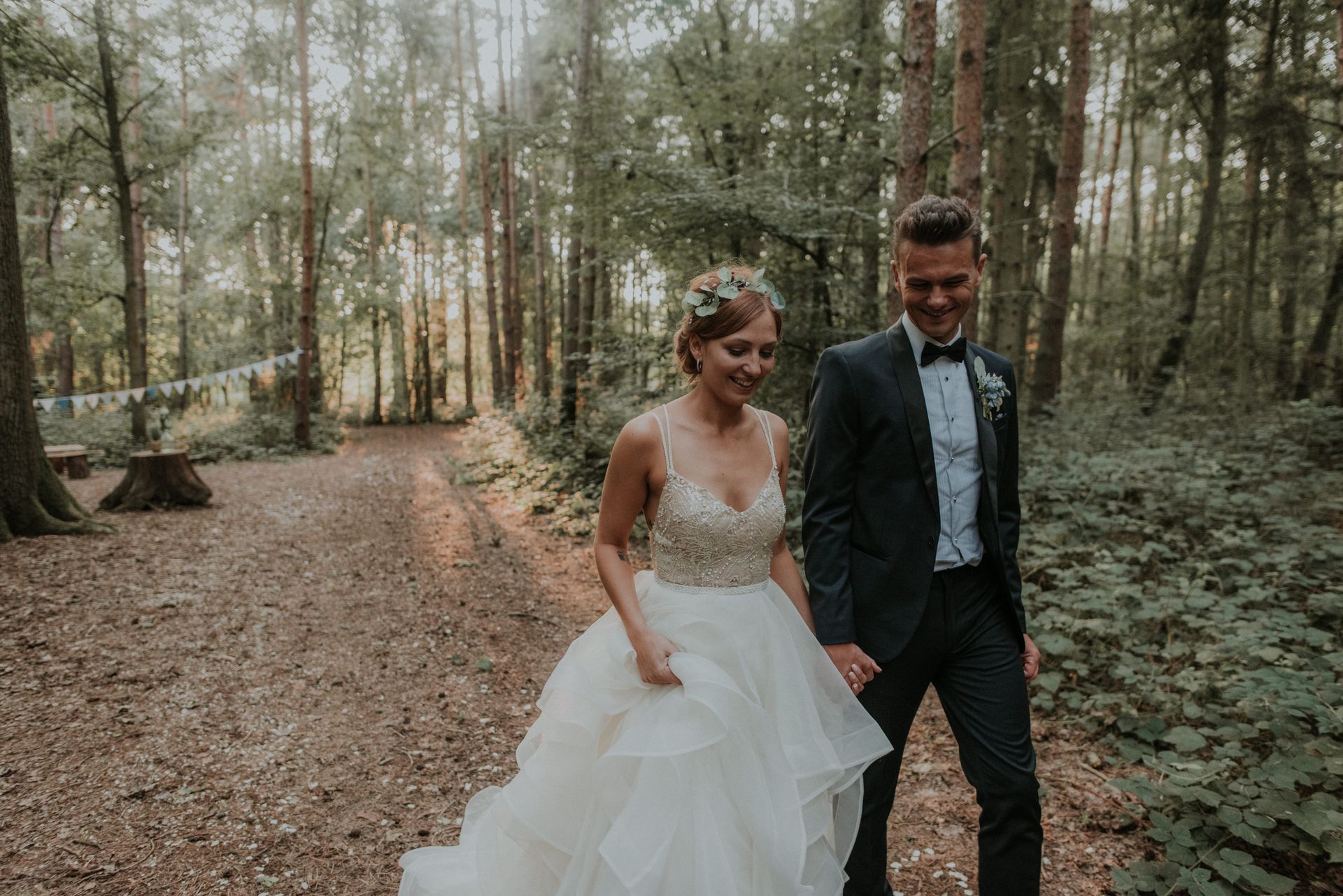 A bride and groom are walking through the woods holding hands.