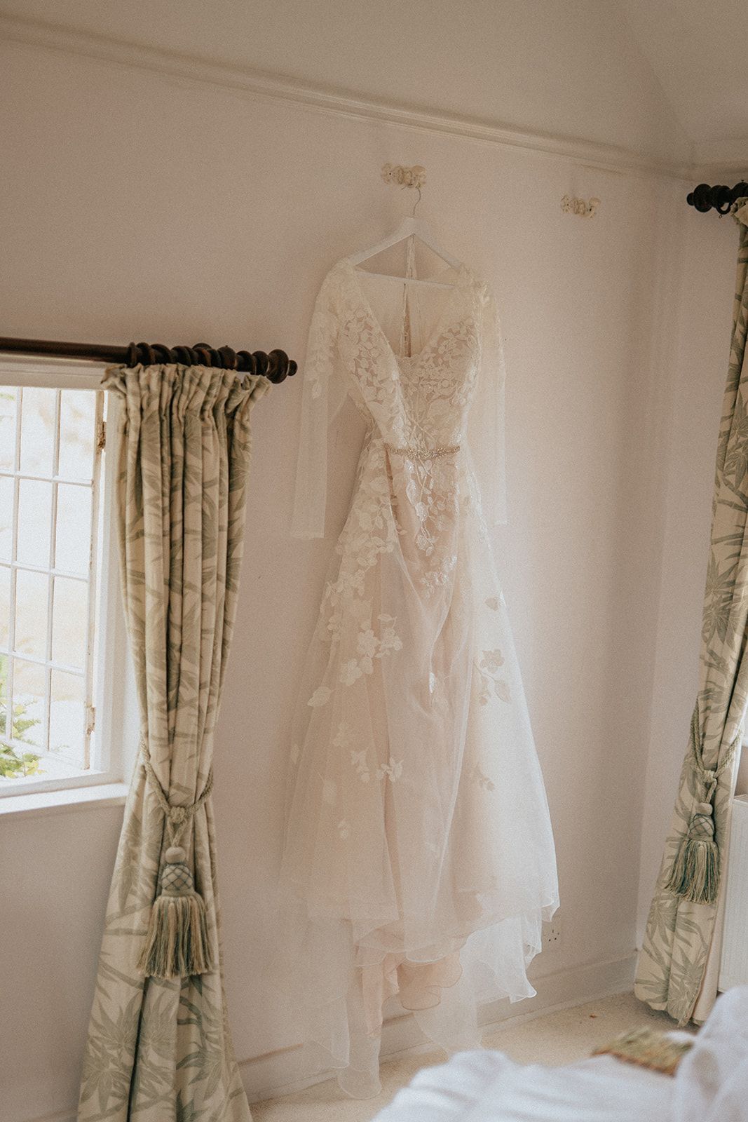 A wedding dress is hanging on a hanger in a room next to a window.