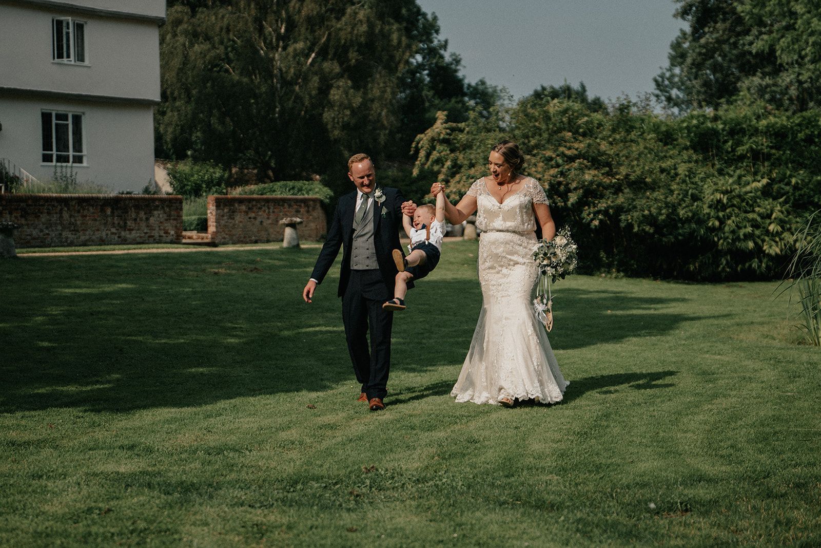 A bride and groom are walking through a grassy field with a child.
