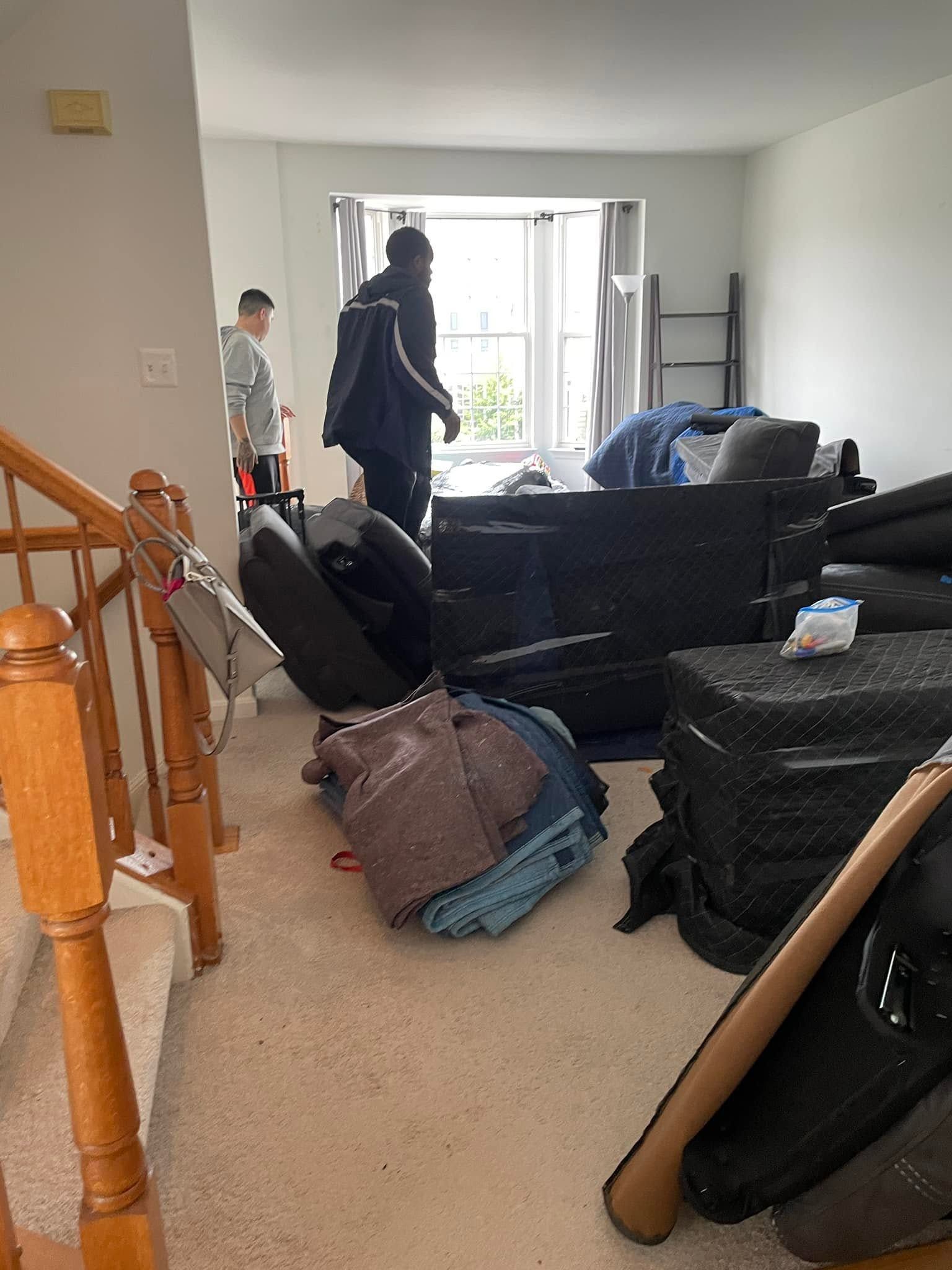 Cluttered living room with stacked furniture and bags; two people stand near a bright window.