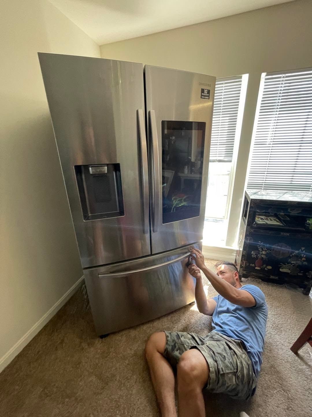 Person kneeling on carpet beside stainless steel refrigerator, reaching toward the lower freezer door.
