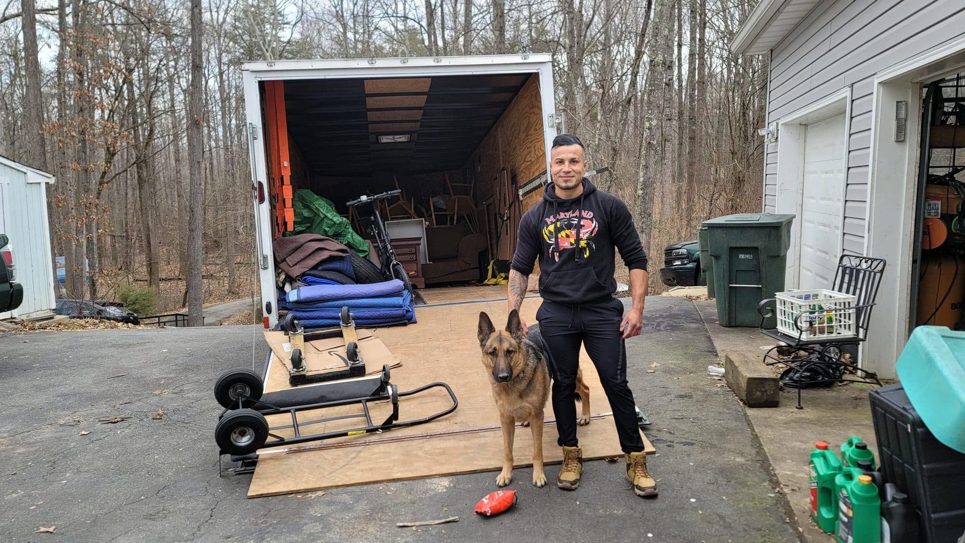 Man standing beside a dog in front of an open moving trailer on a driveway