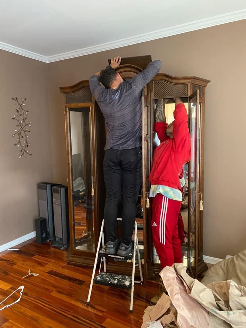 Two people assembling a tall wooden wardrobe in a room, one on a step ladder and one standing beside it.
