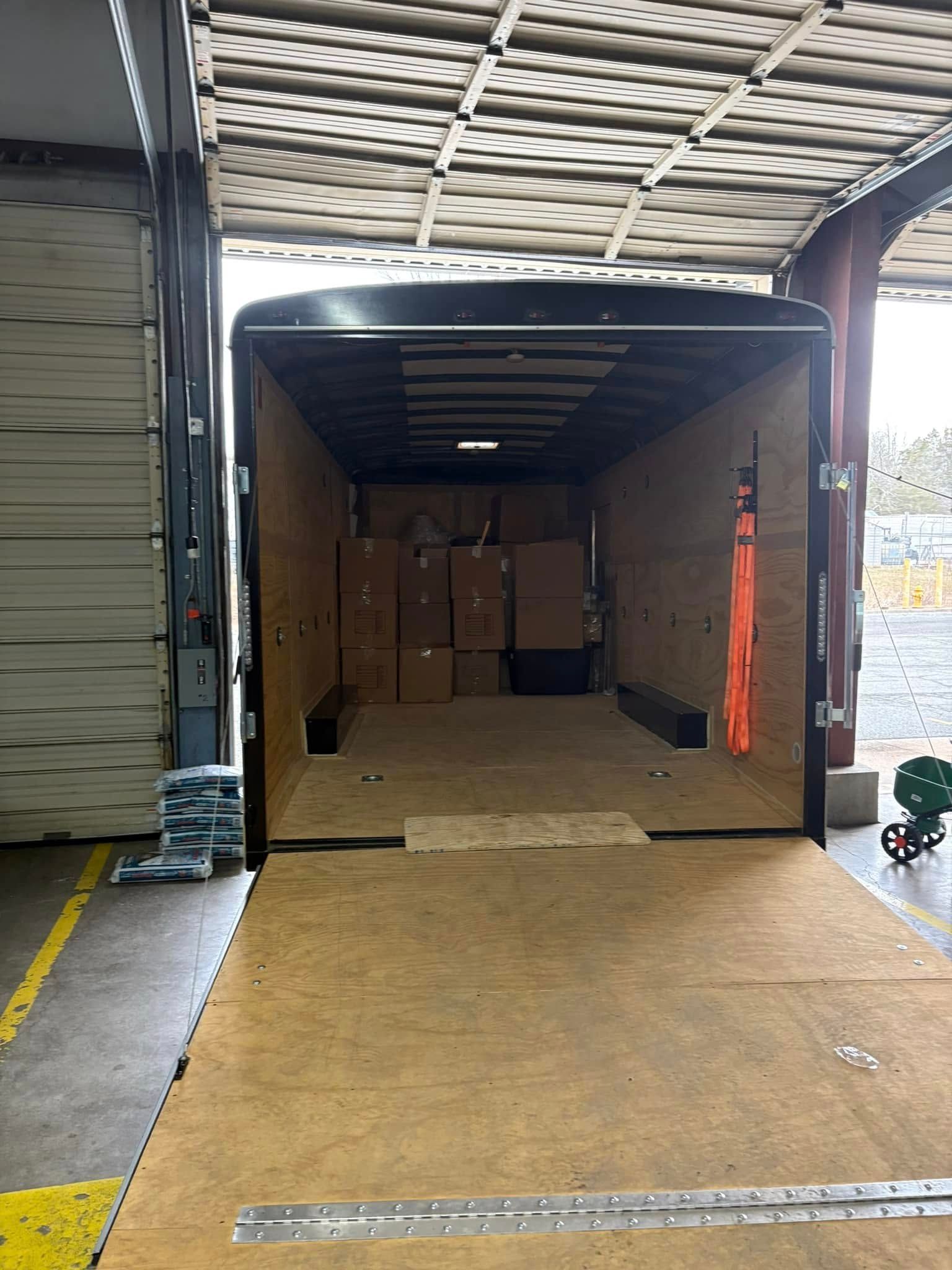 Open cargo trailer loaded with stacked cardboard boxes, backed into a warehouse bay.