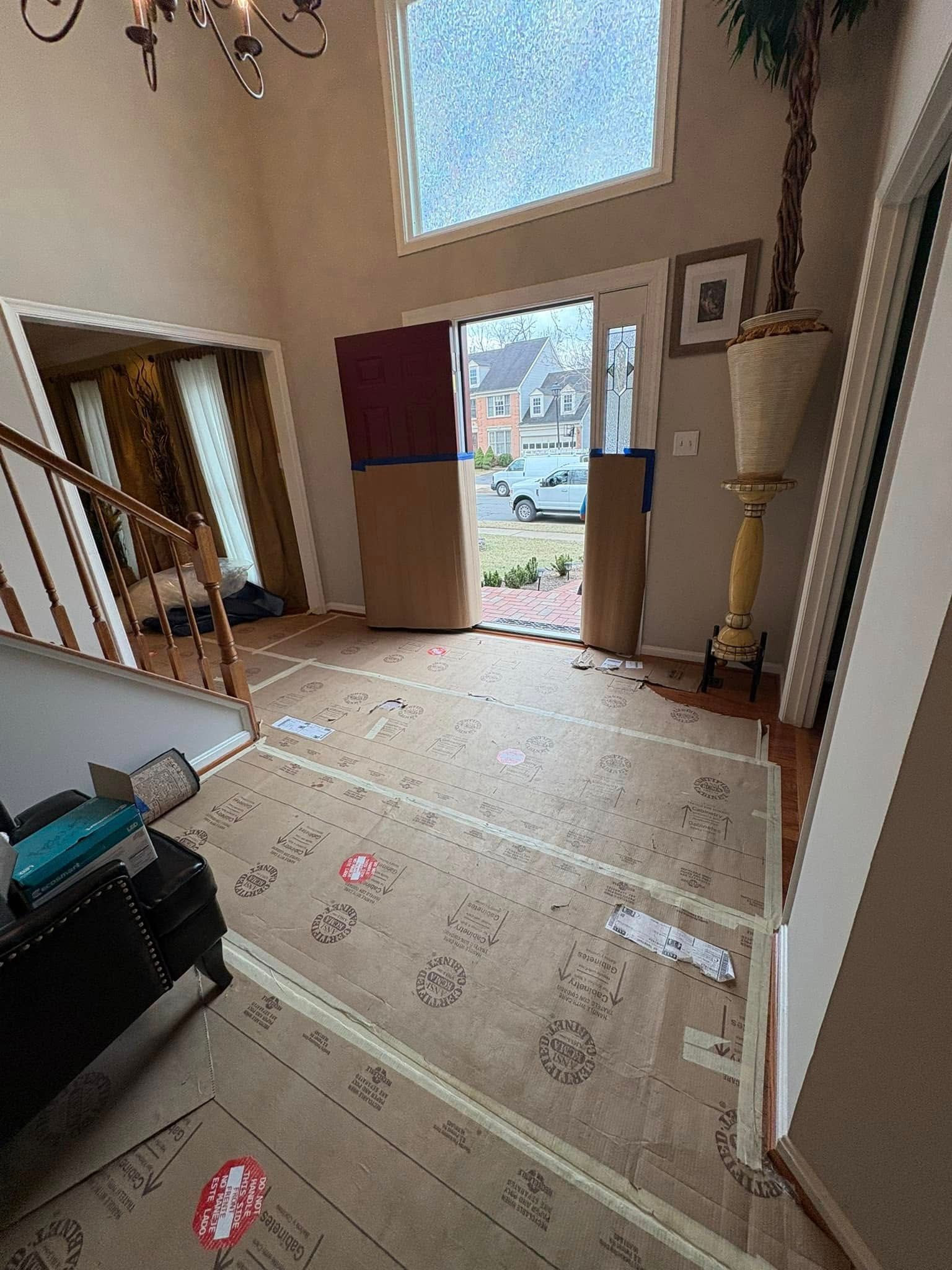 Messy entryway with cardboard floor protection, open front door, and stairs leading down to the left.