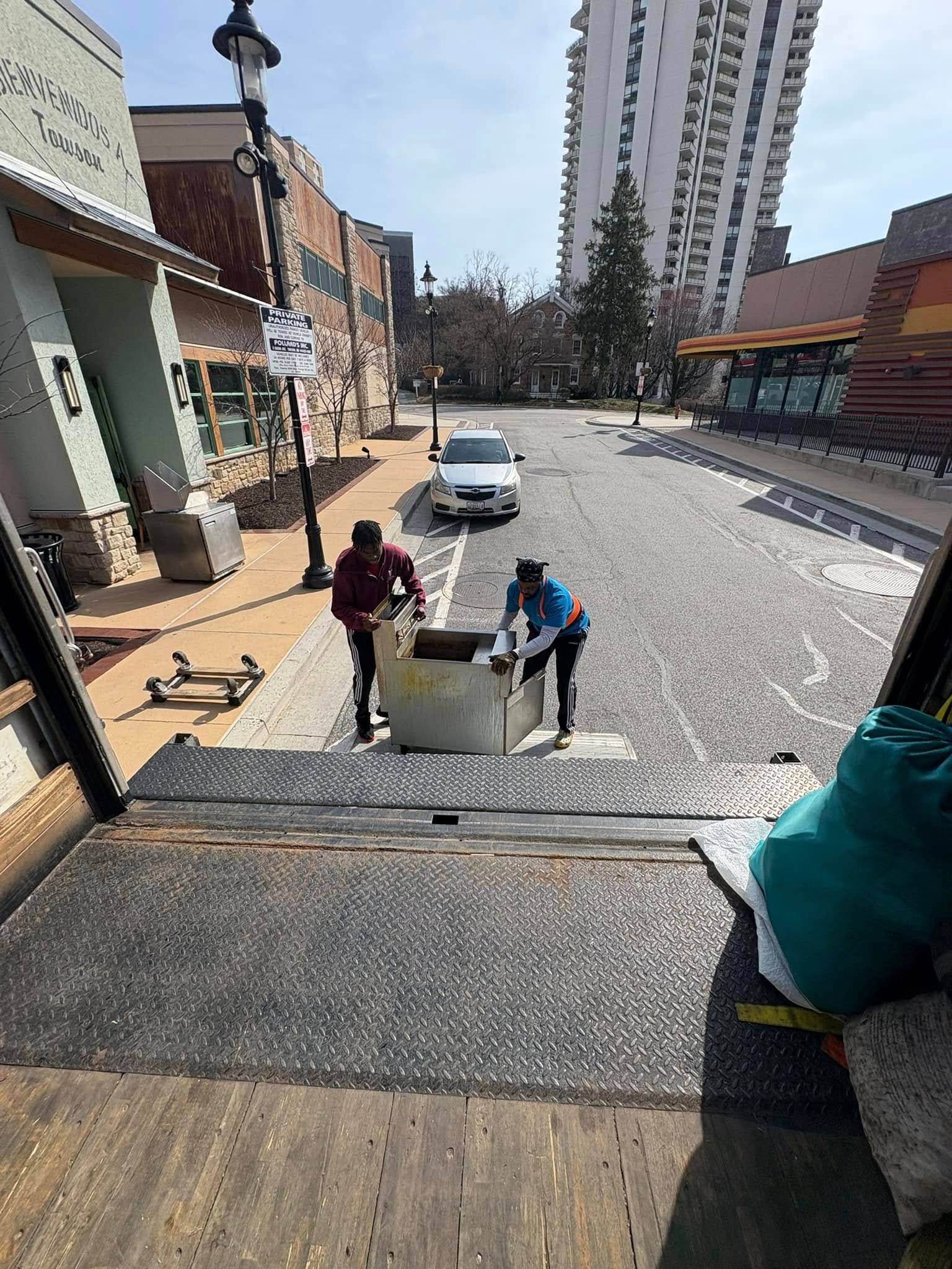 Two workers push a concrete barrier down a cobblestone street with buildings and a car nearby.