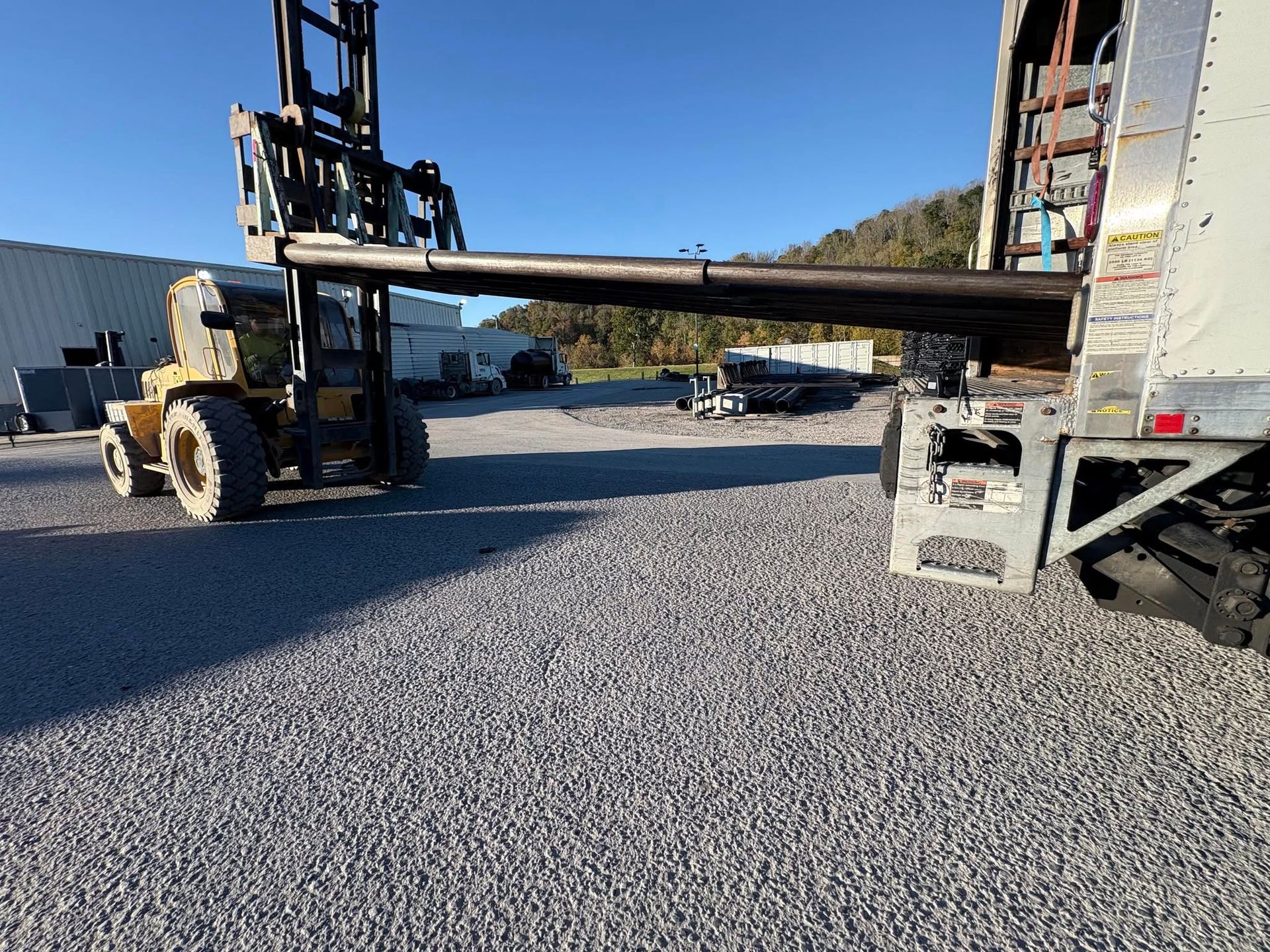 Forklift loading a long steel beam onto a flatbed truck in a gravel yard.